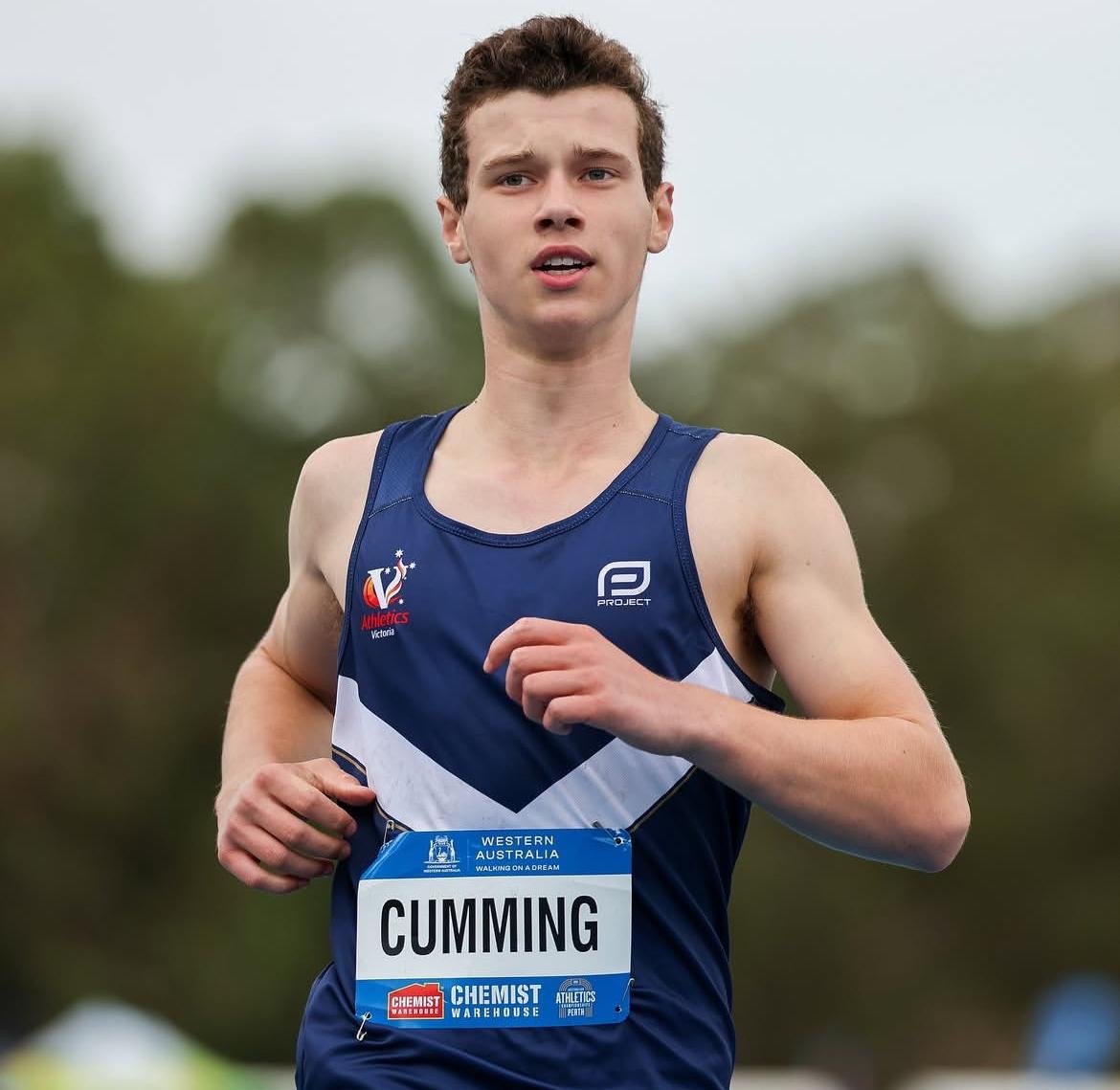 Young male cross-country runner wearing a blue racing uniform with a bib labeled 'CUMMING' during a race outdoors.