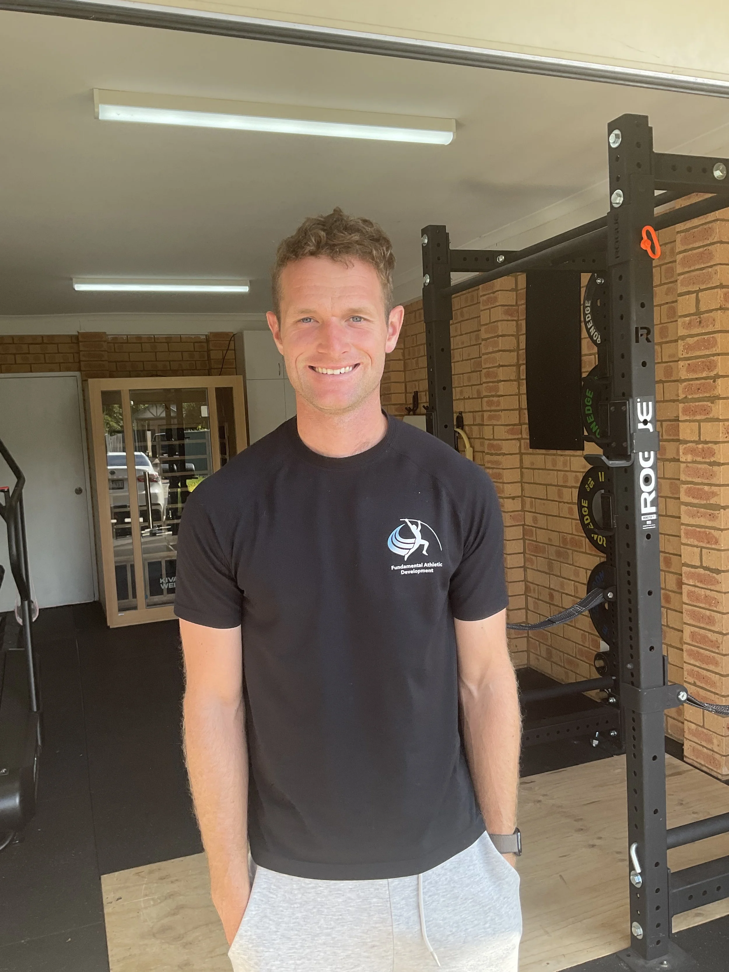 A smiling man with curly hair wearing a black t-shirt and gray shorts standing inside a gym next to a black workout rack.