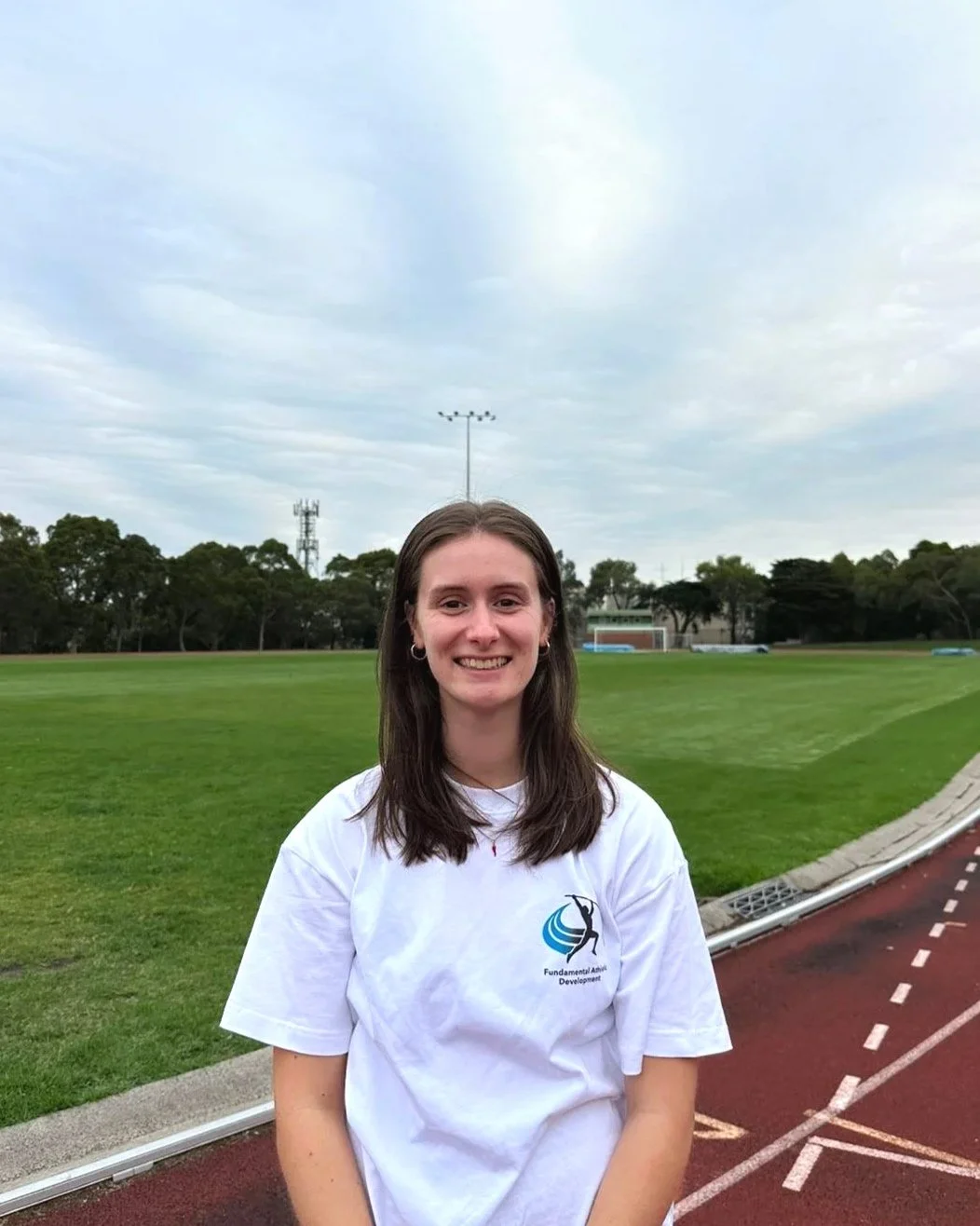A young woman smiling outdoors on a track and field with green grass and trees in the background, wearing a white T-shirt with a logo reading "Fundamental Athletic Development."