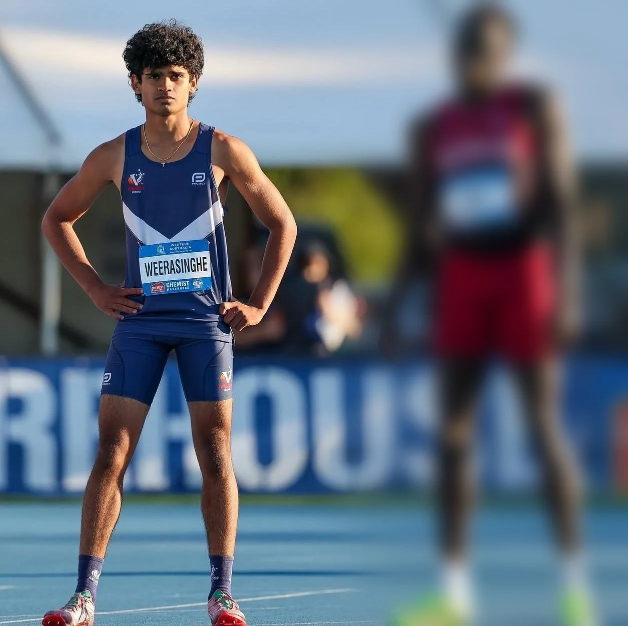 A young male athlete in a blue running uniform standing on a track with hands on hips, looking ahead, with a blurred runner in red and black in the background during a daytime outdoor race.