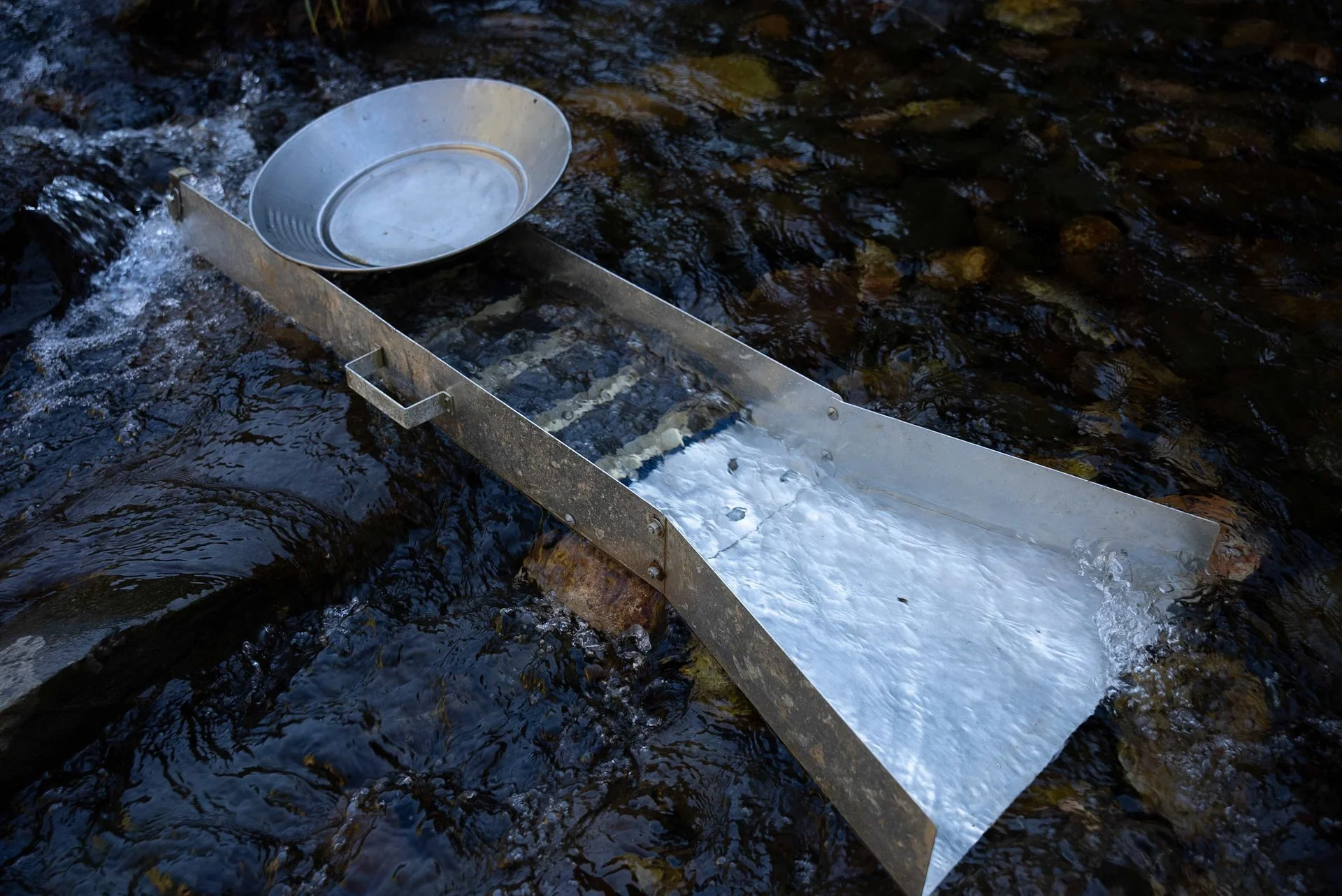 A metal stream sampling device with a round dish on top, floating on a river with dark water and rocks.