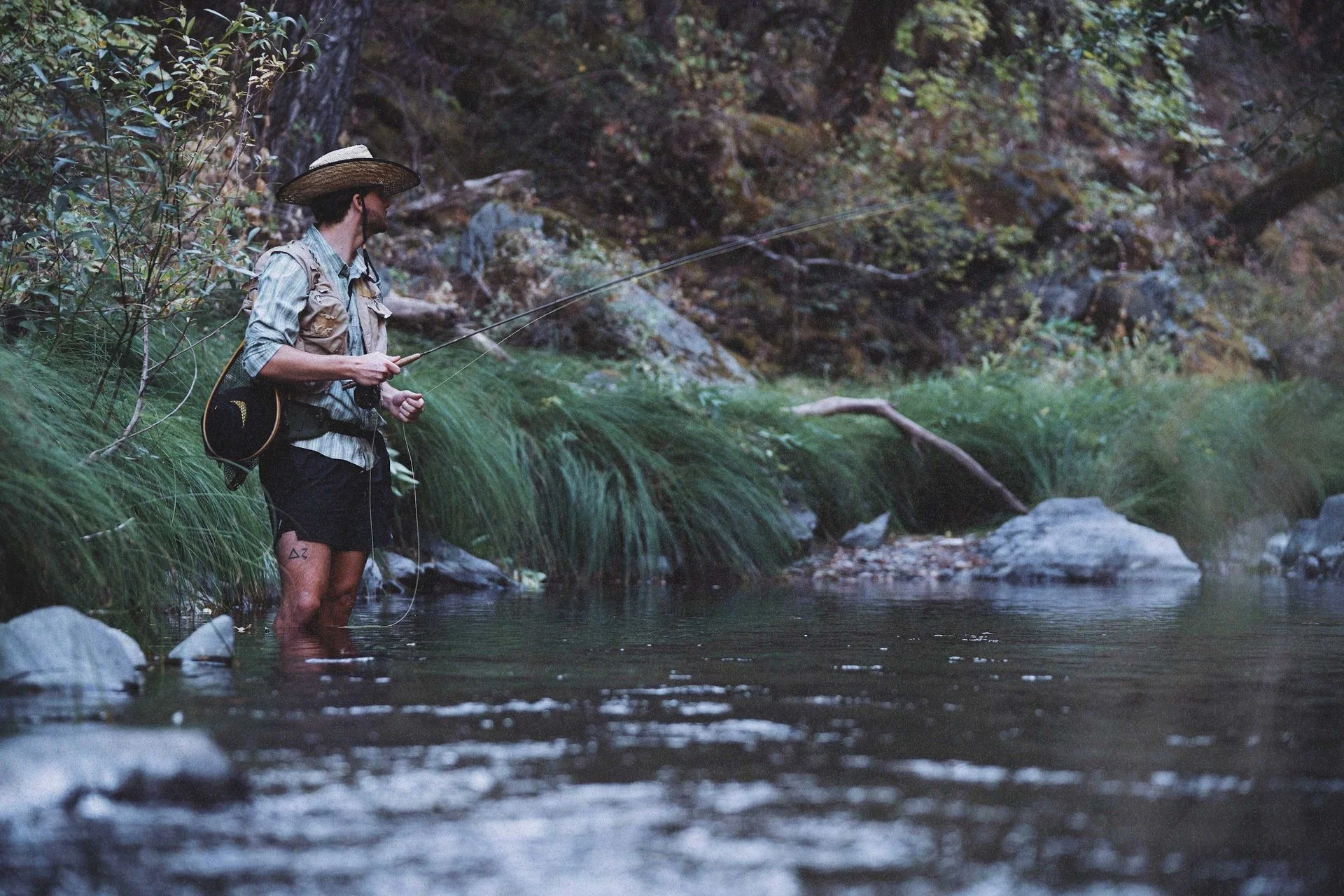 A man fishing in a river in a forest, wearing a wide-brimmed hat, a fishing vest, shorts, and standing in the water.