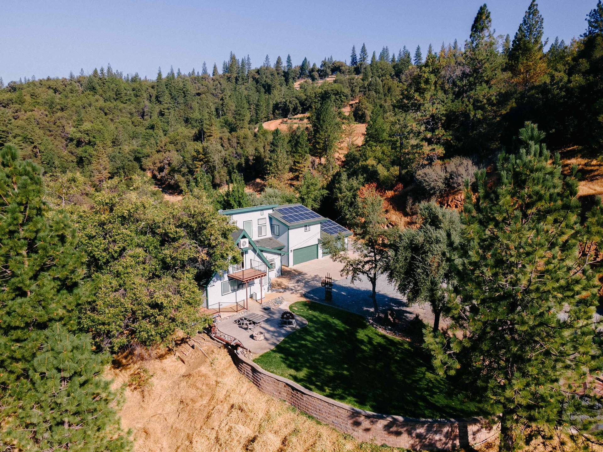 A modern house with solar panels on the roof, surrounded by trees and a grassy yard, in a hilly forested area.