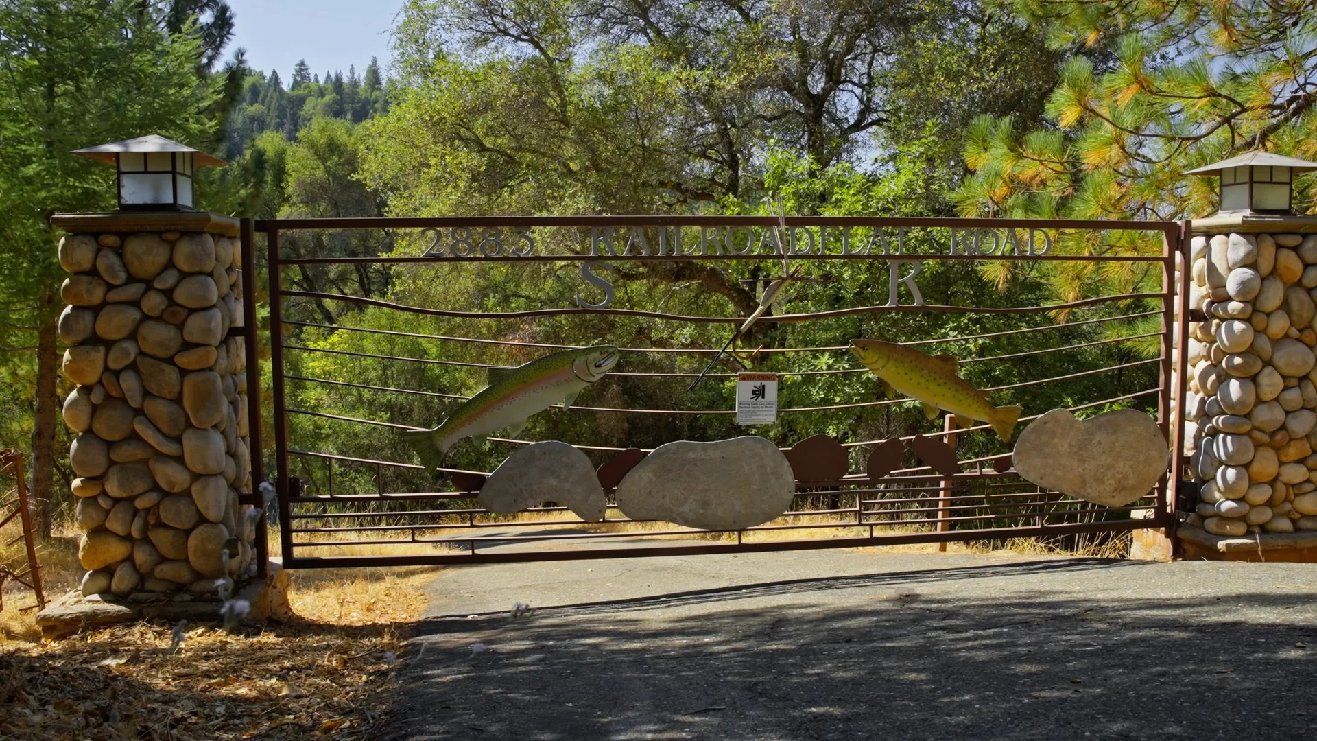 A wrought iron gate with stone pillars on each side, featuring two large fish sculptures and decorative rocks attached, leading to a wooded area with trees and mountains in the background.