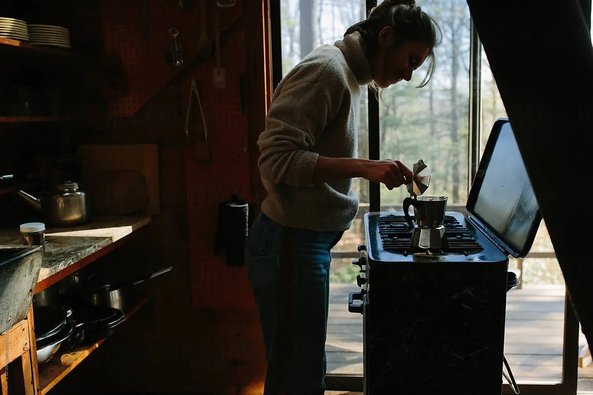 Woman preparing coffee on a small stove in a cozy kitchen next to a large window