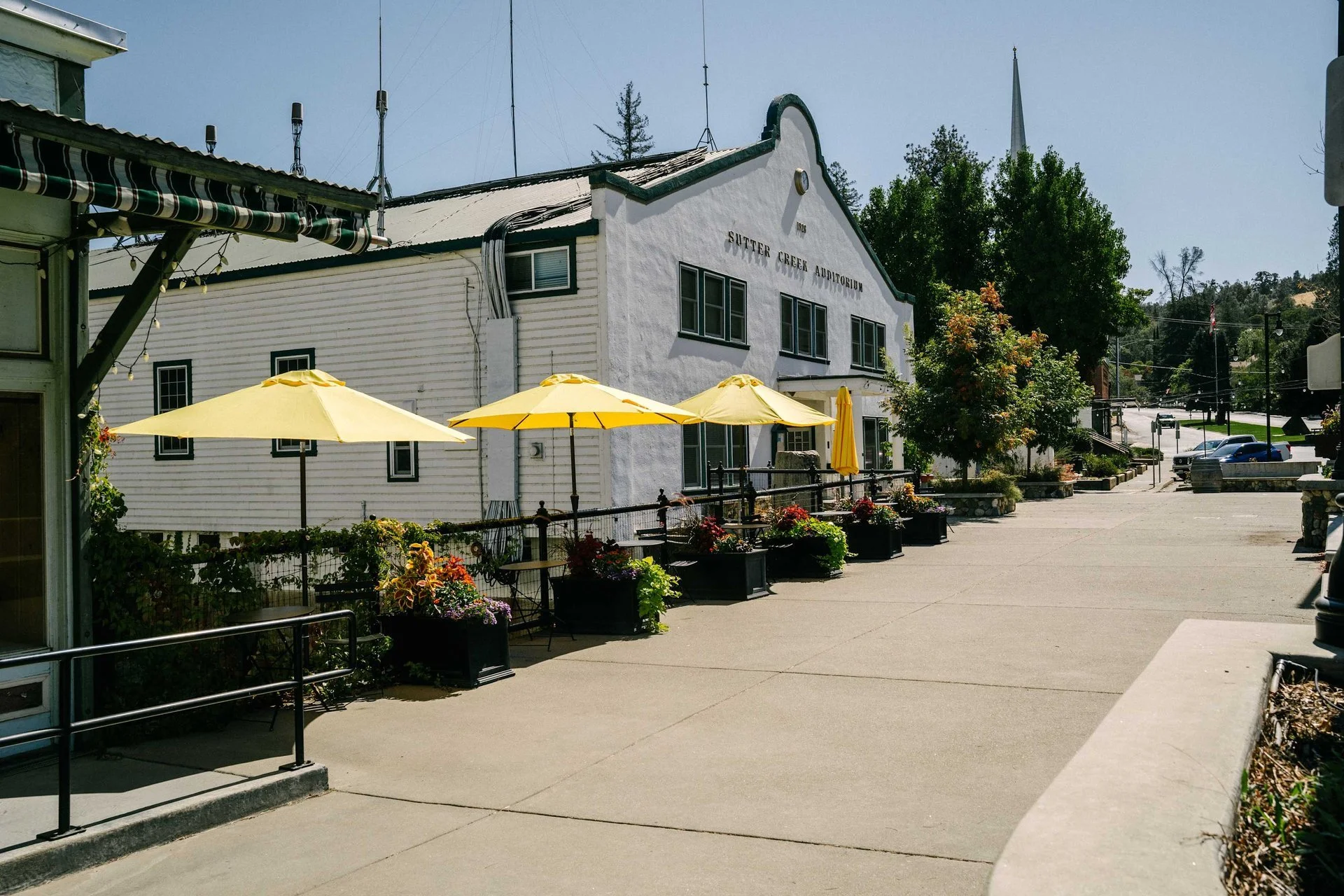 Empty outdoor patio with yellow umbrellas, flower planters, and a building labeled 'Sutter Creek Auditorium' in a small town setting.