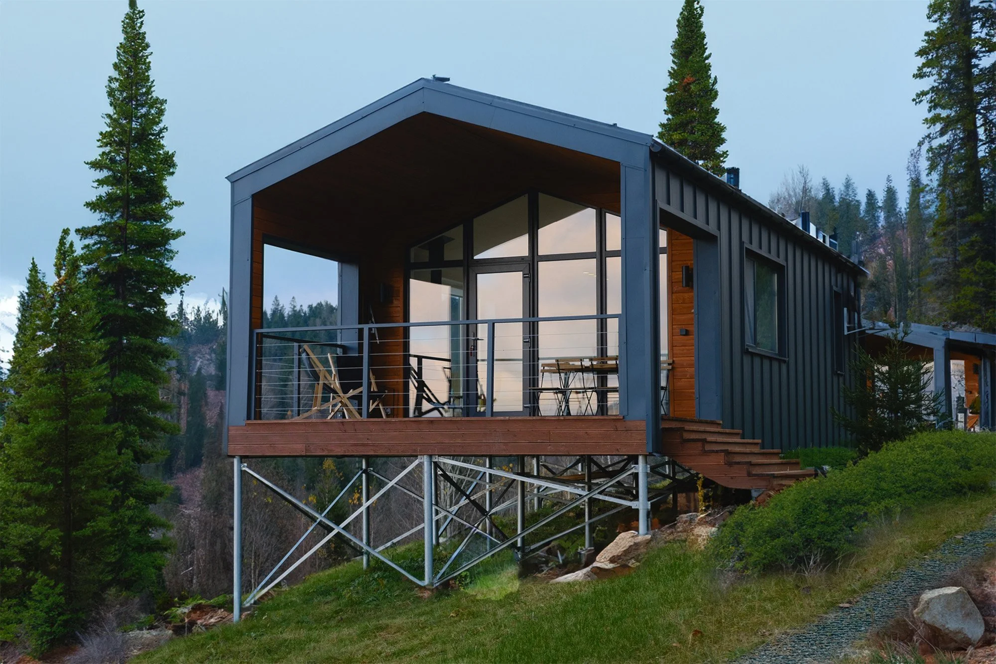 Modern house built on stilts on a grassy hillside surrounded by trees, with large glass windows and a wooden balcony.