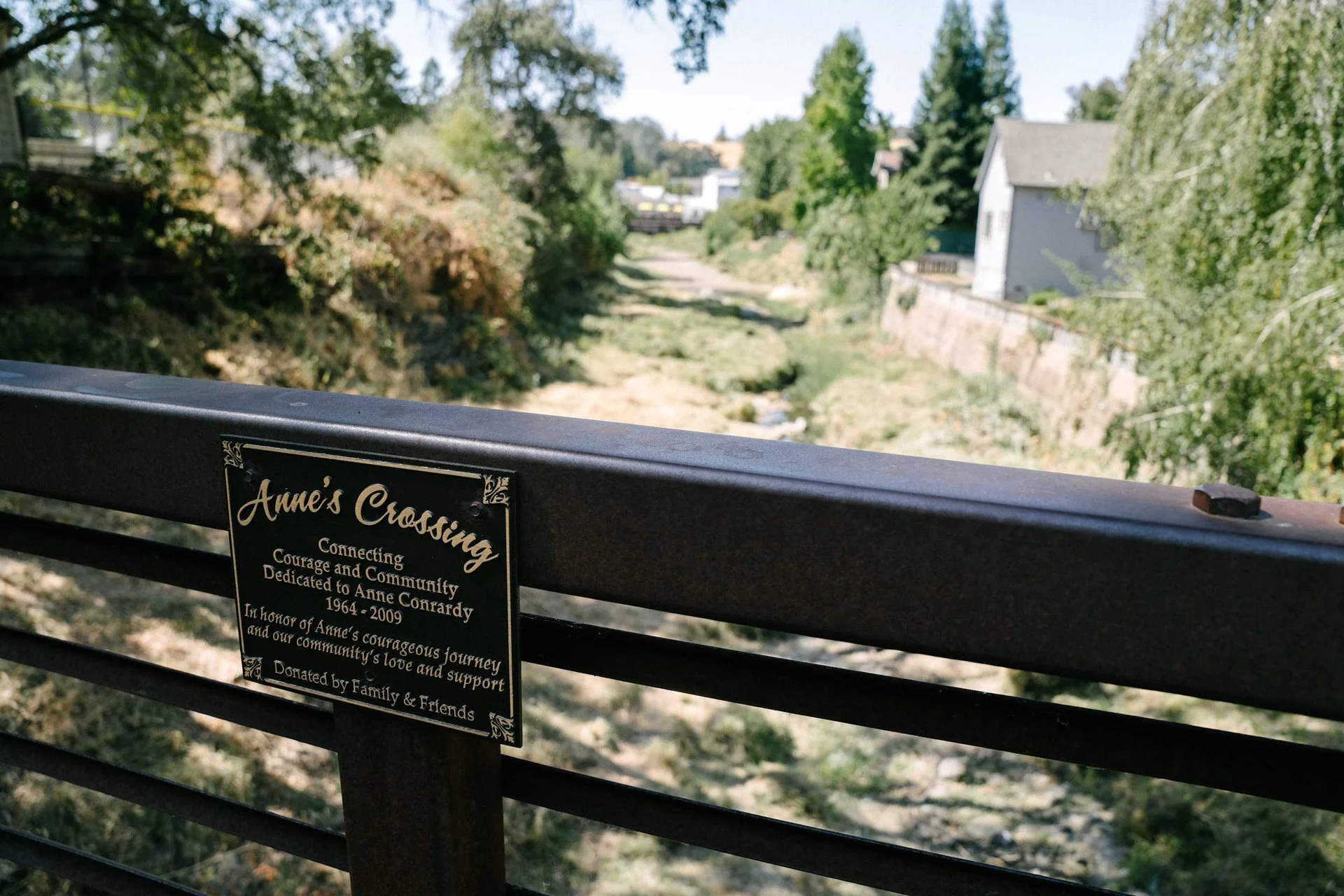 A bridge over a dry creek bed with a plaque that reads 'Anne's Crossing, Connecting Courage and Community, Dedicated to Anne Conardy 1964-2009, In honor of Anne's courageous journey and our community's love and support, Donated by Family & Friends.'