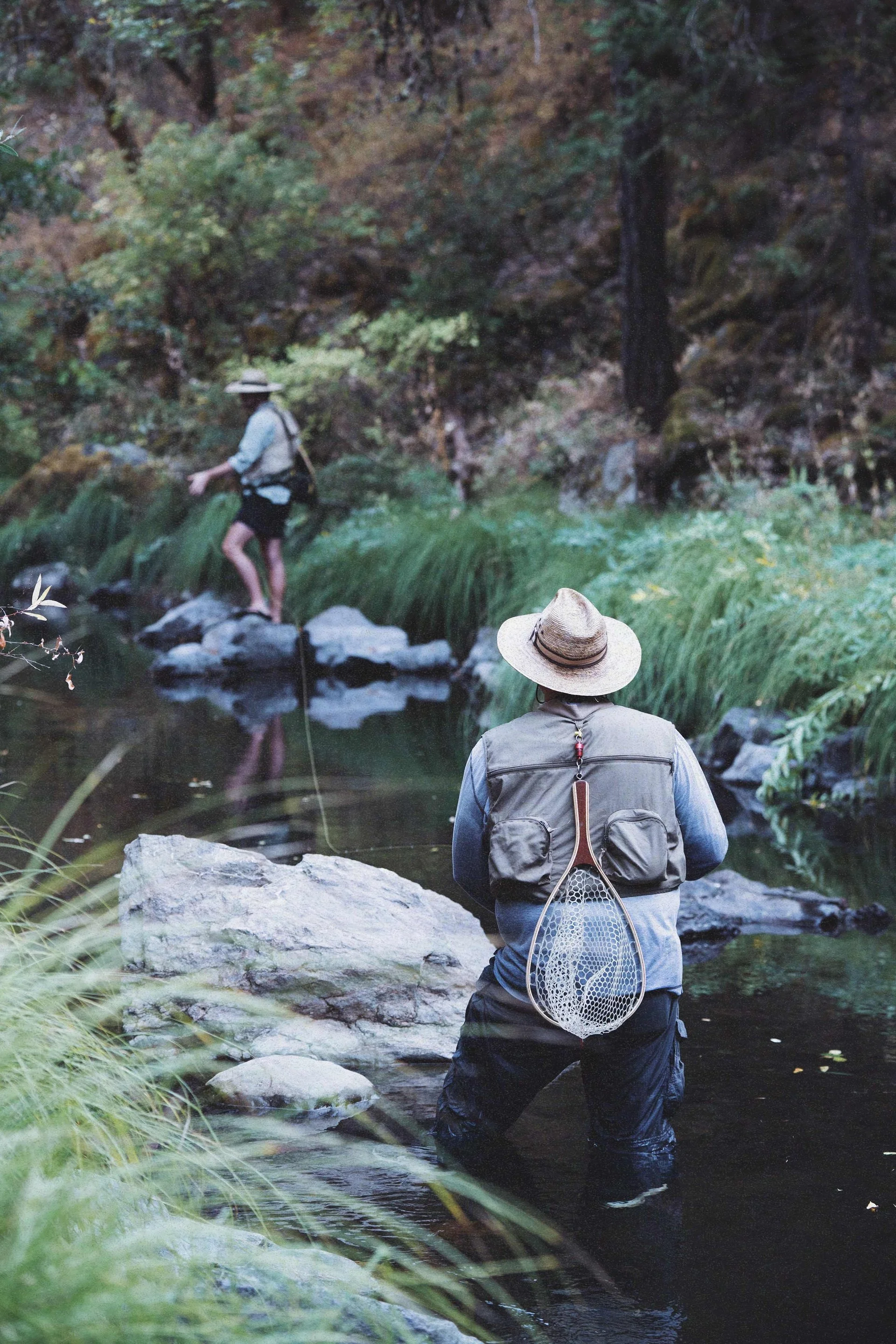 Two people fishing in a stream in a forested area with trees and rocks. One person is in the foreground wearing a hat, vest, and waders, holding a fishing net, while the other person is in the background on rocks.