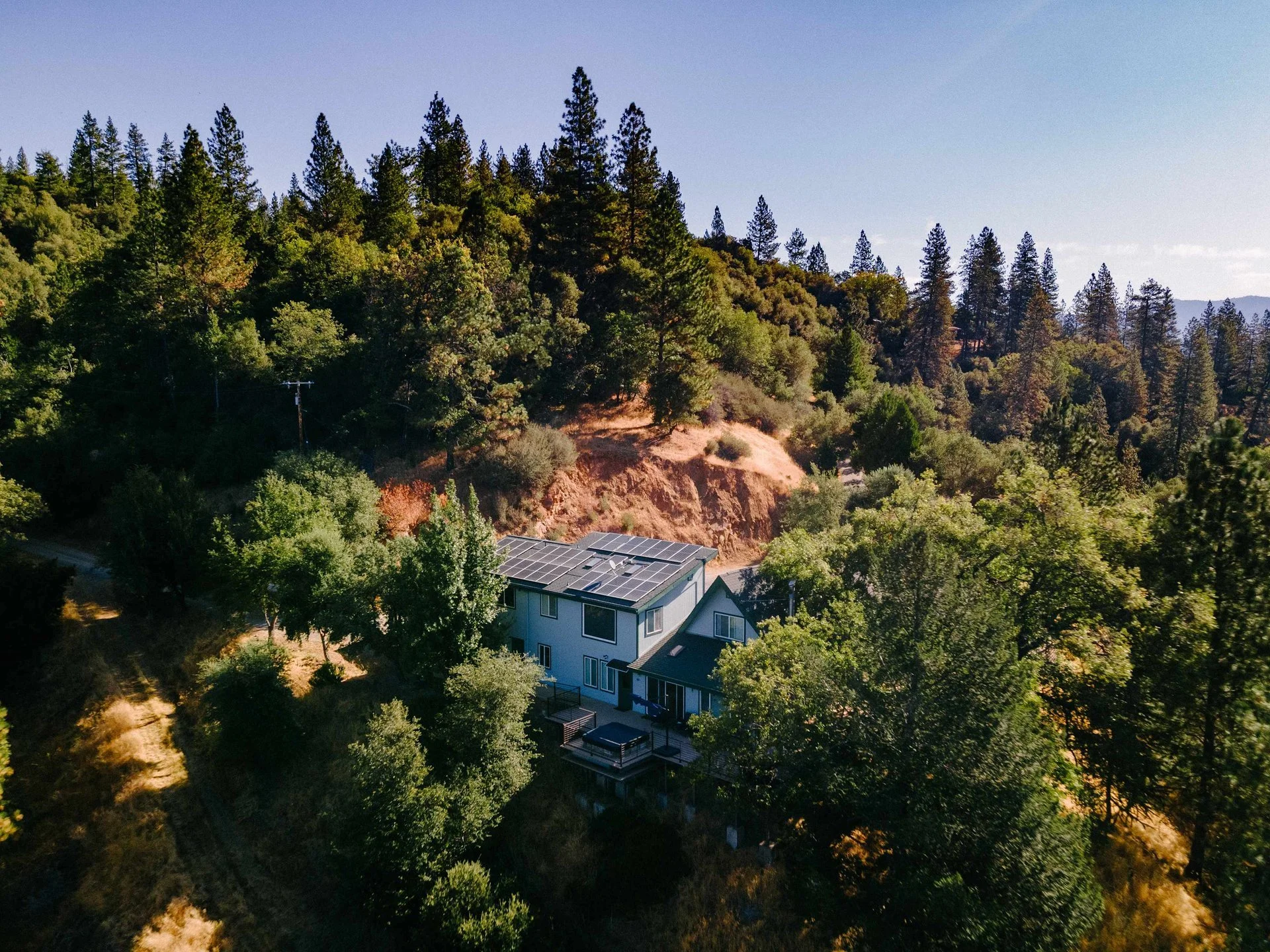 A house with solar panels on the roof surrounded by trees on a hillside, set against a clear blue sky.