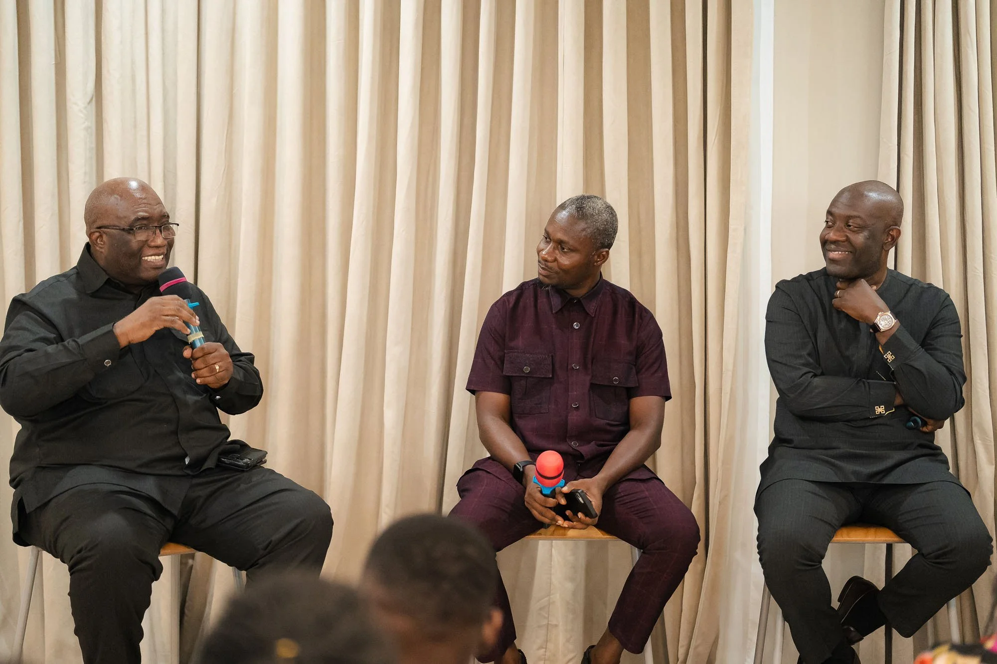 Three men seated on a stage in front of beige curtains, engaged in a discussion. The man on the left holds a microphone and speaks, while the other two listen attentively.