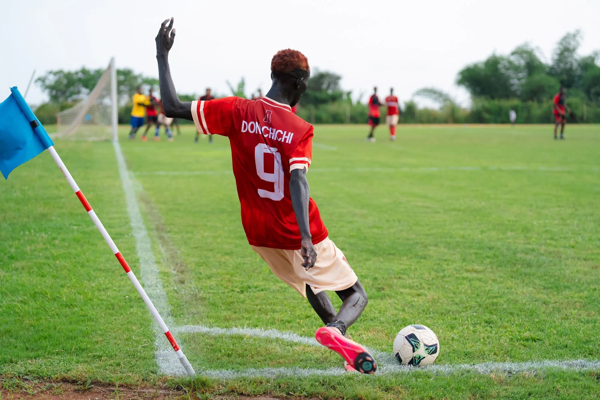 Soccer player in red jersey kicking a ball near the corner flag during a match on a grassy field with other players in the background.