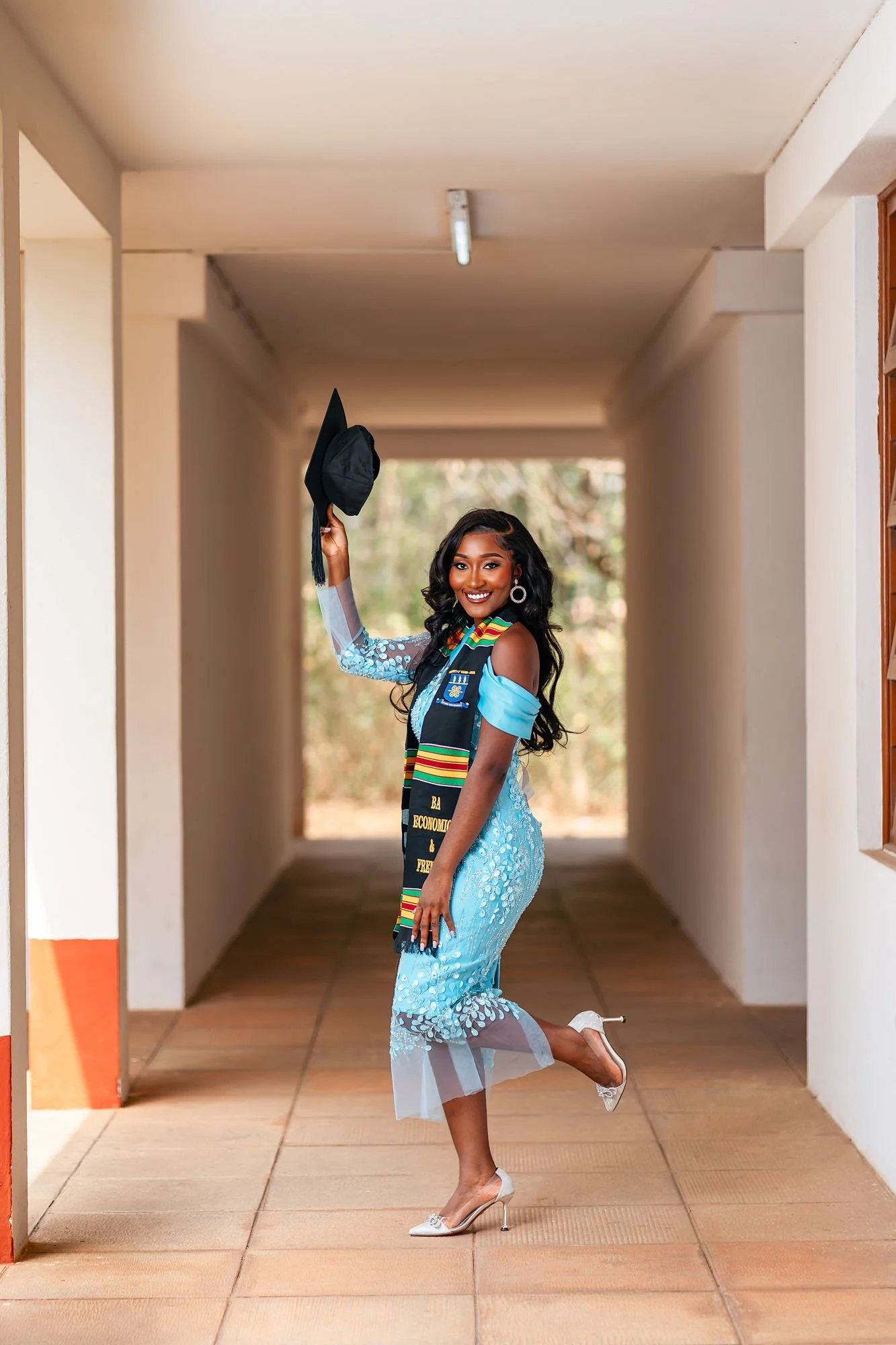 A young woman in a blue dress with floral details celebrating her graduation, holding her cap in the air and smiling, standing in an outdoor corridor.
