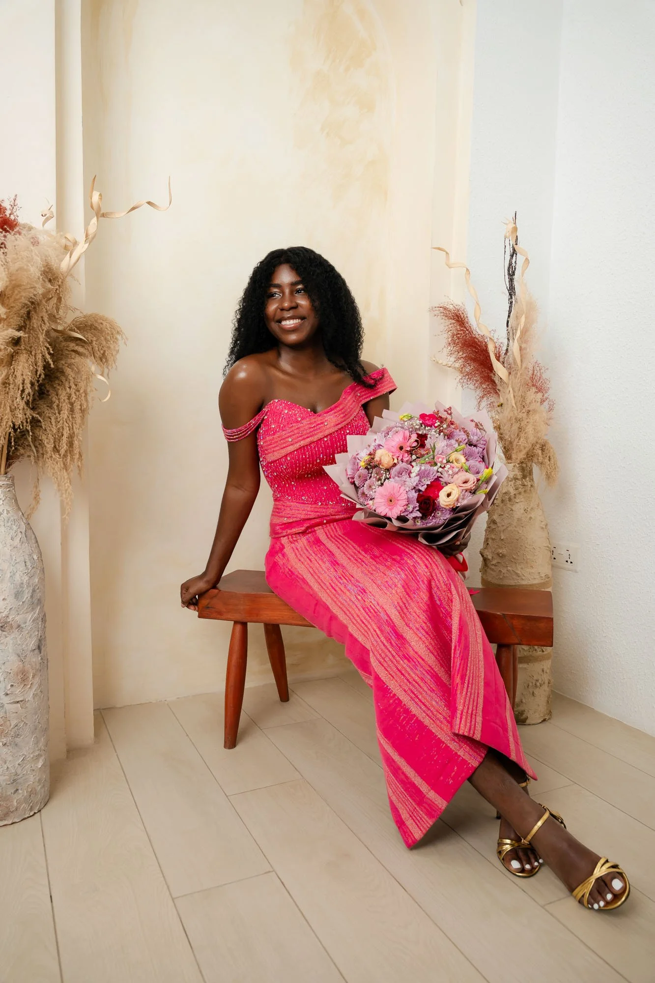 A woman in a pink dress sitting on a wooden bench, holding a bouquet of pink and purple flowers, smiling in a decorated room with beige and brown dried plants.