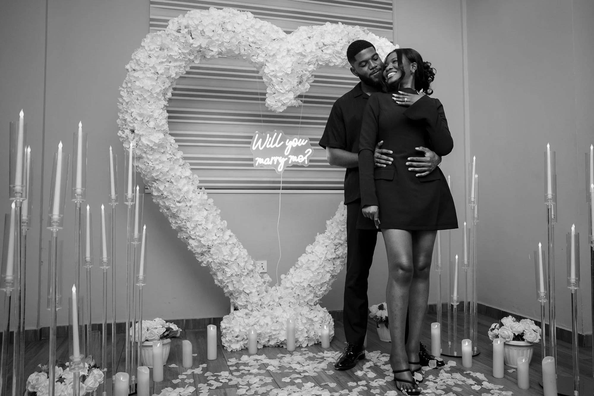 A couple is embracing in front of a decorative heart-shaped floral arrangement and candles, with a neon sign reading 'Will you marry me?' behind them. The woman is smiling, and the man has his hands on her waist.