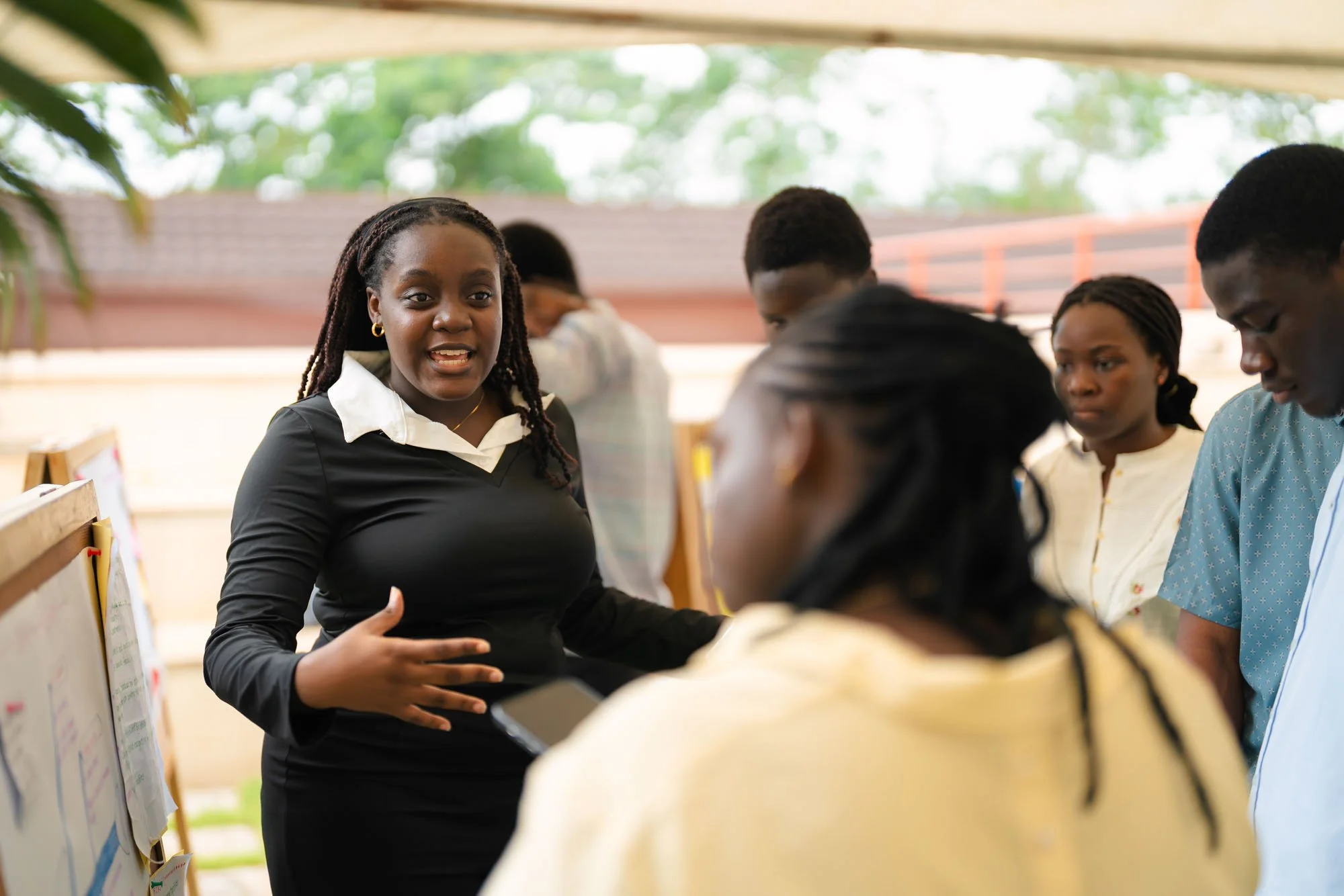 A woman in a black dress is speaking to a group of people in an outdoor setting, with trees and a wooden fence in the background.