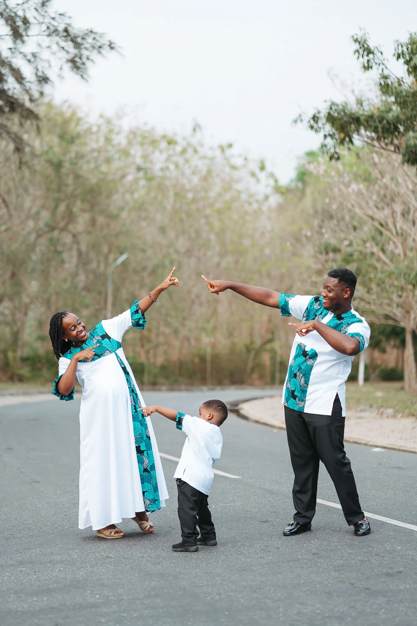 A family of three, a woman, a man, and a young boy, dancing and smiling on an empty street lined with trees, all wearing matching African print shirts.