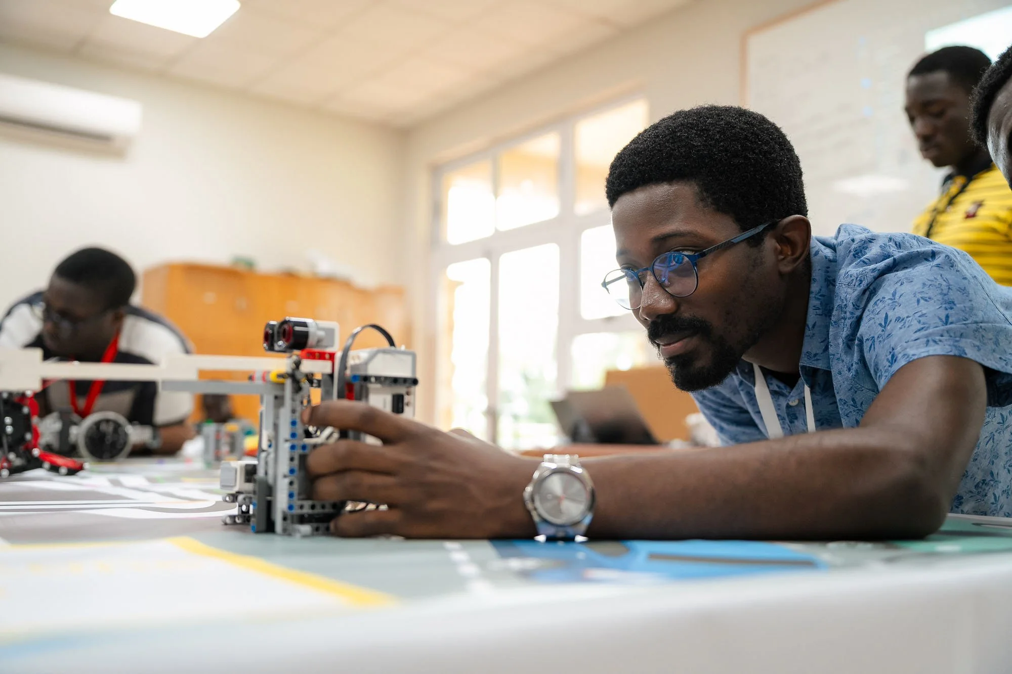 A man with glasses and a blue patterned shirt working on a robotics project at a table in a well-lit room with large windows, with two other people in the background.