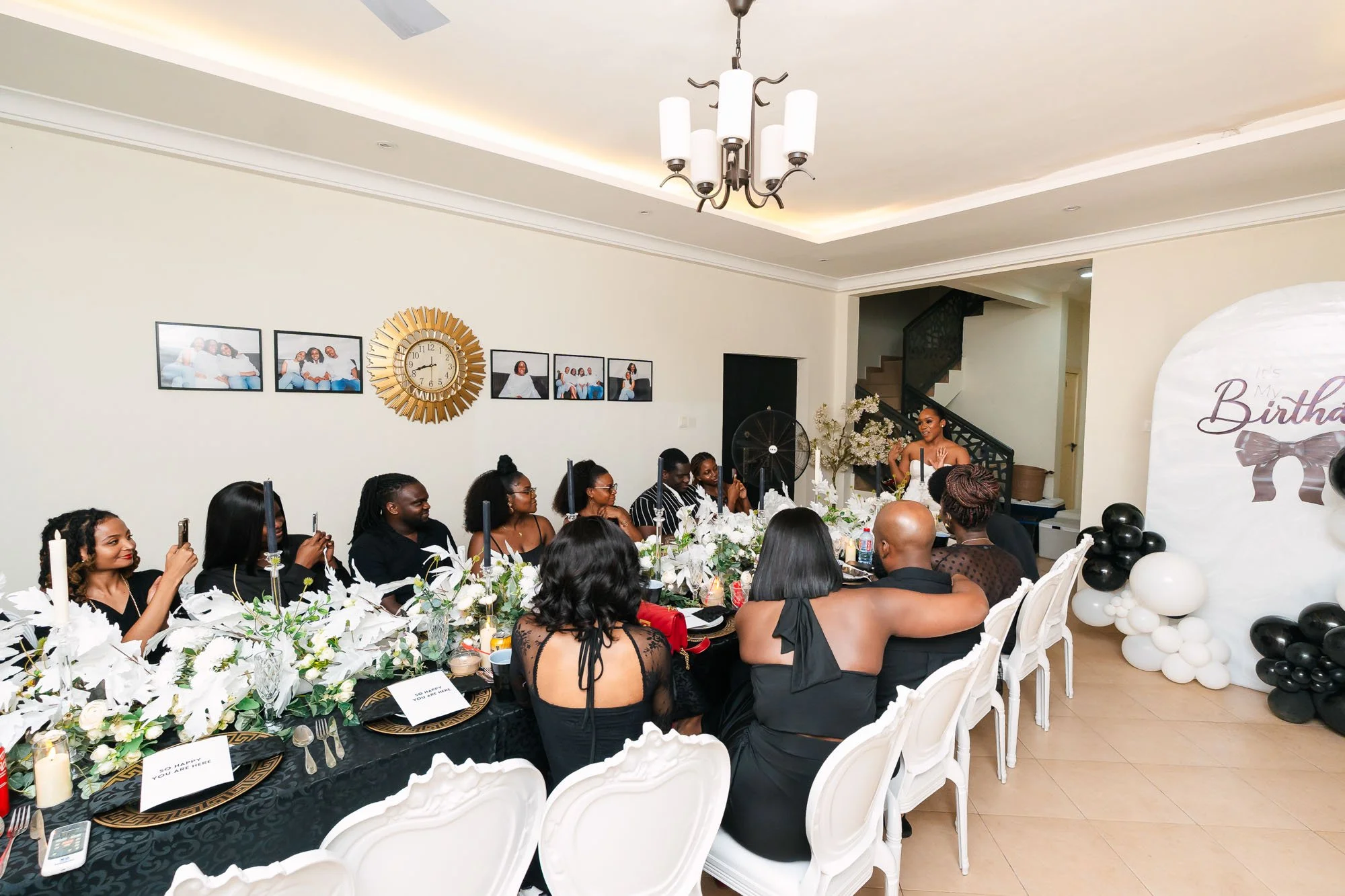 A birthday celebration with a group of people sitting at a decorated table, a woman speaking at a microphone near a backdrop with balloons and a 'Happy Birthday' sign, in a well-lit room with wall art and a staircase.