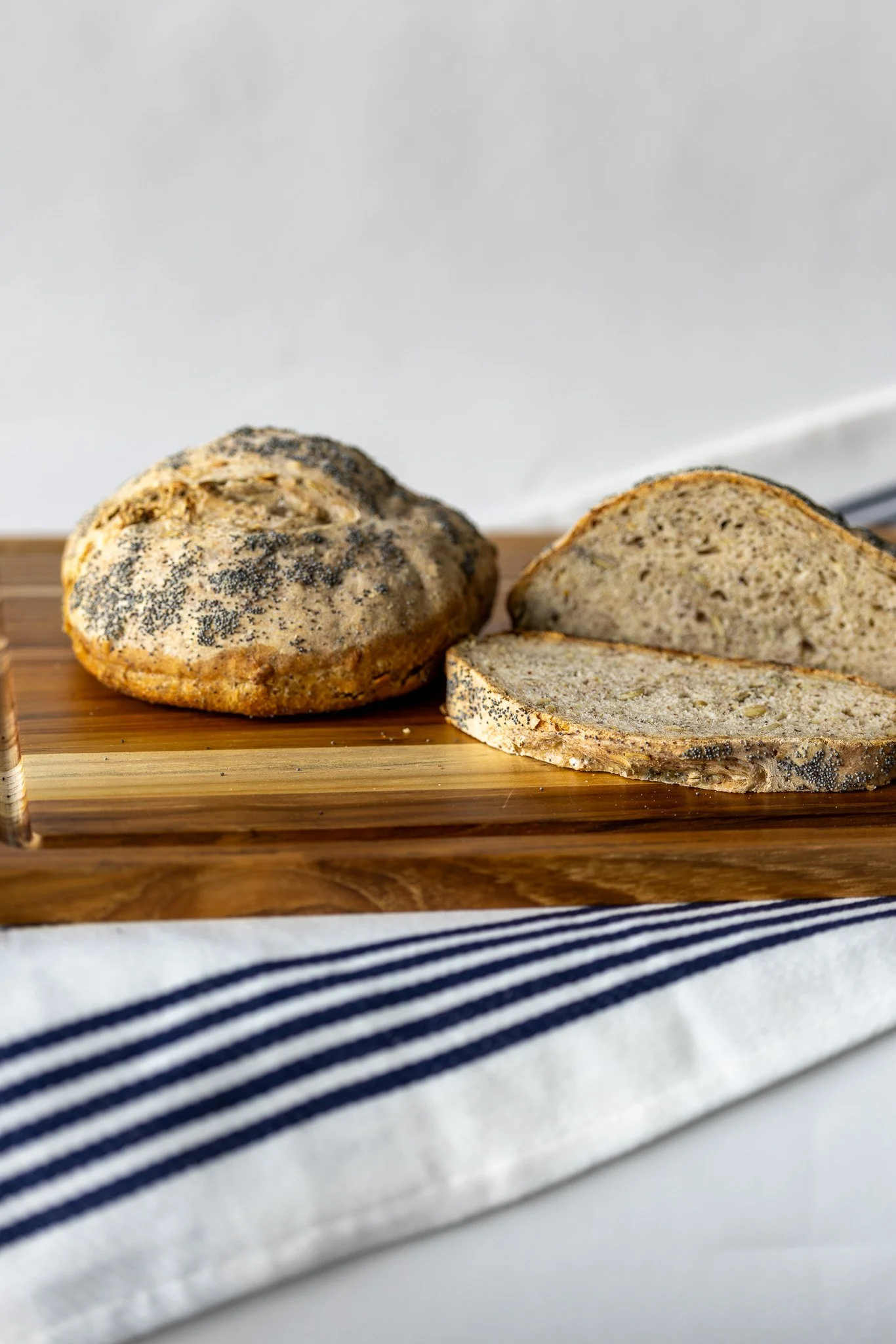 Close-up of Arctic Sourdough sliced multigrain bread with poppy seeds on a wooden cutting board, placed on a white and navy striped cloth.