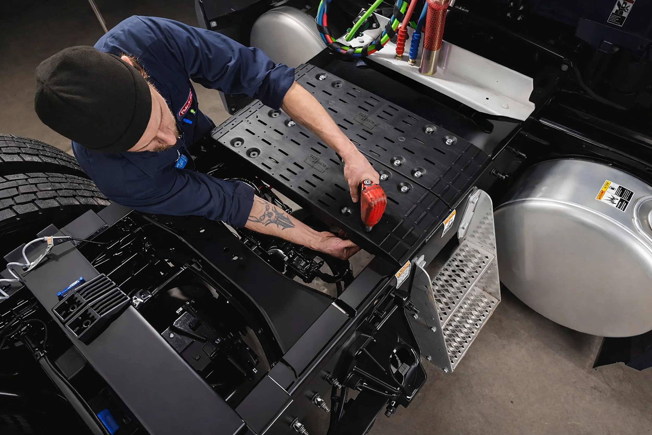 A worker installing or repairing components on a semi-truck chassis, using a power tool, with the truck's metal frame and various parts visible.