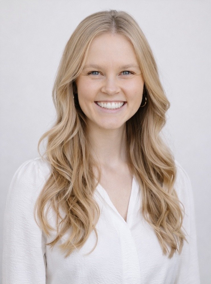A woman with long, wavy blonde hair, blue eyes, and a bright smile, wearing a white blouse, standing against a plain, light-colored background.