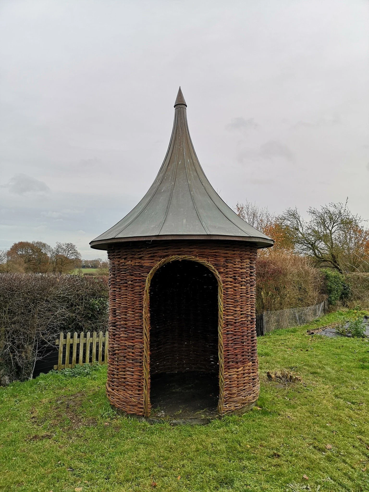 Wicker gazebo with conical metal roof on a grassy area, trees and a cloudy sky in the background.
