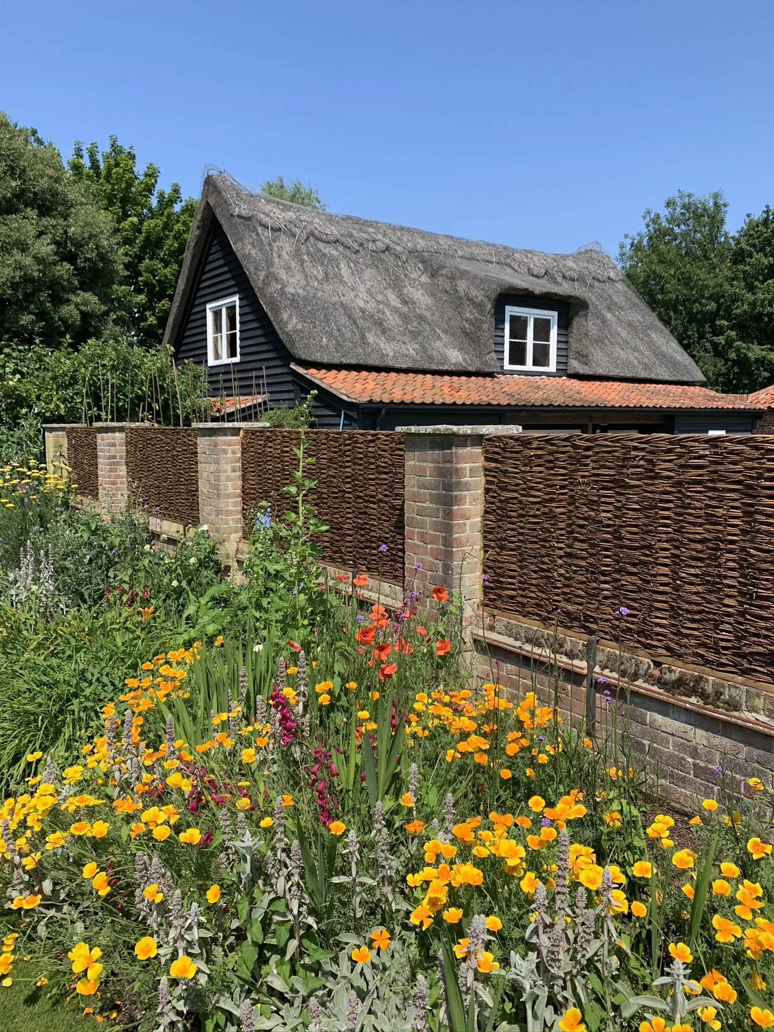 A house with a thatched roof, black wooden siding, and white-framed windows, surrounded by a garden with colorful flowers and a woven fence, under a clear blue sky.