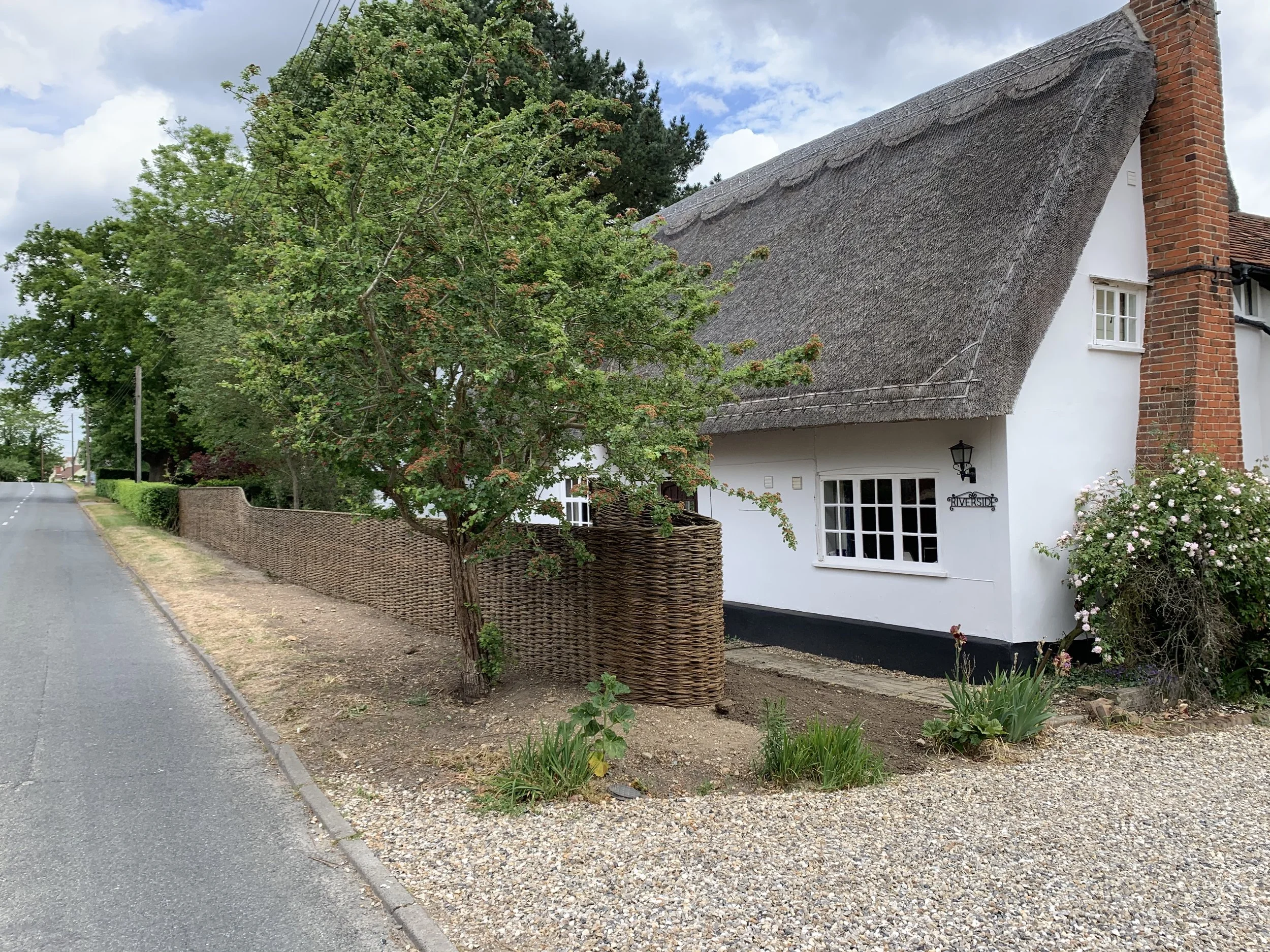 A white cottage with a thatched roof and a brick chimney, surrounded by a woven wooden fence and garden plants, along a quiet roadside.