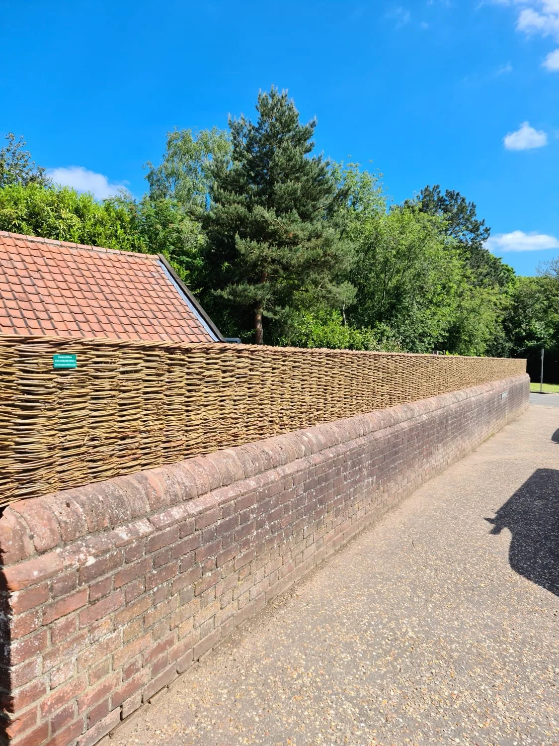 A brick wall with a woven wooden fence on top, green trees, and a blue sky with some clouds.