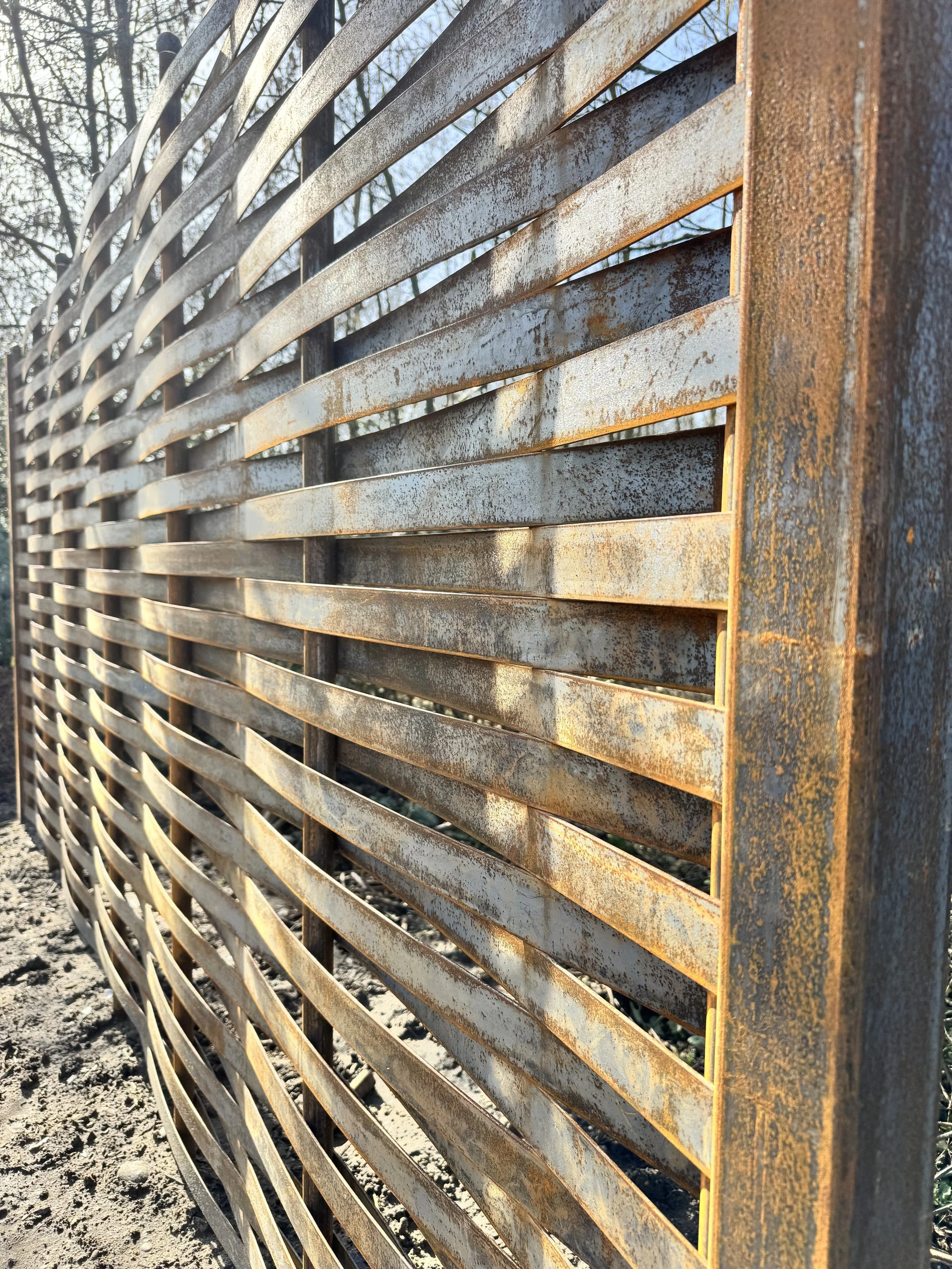 Close-up of a weathered, rusty metal fence with horizontal slats, with trees and blue sky visible in the background.