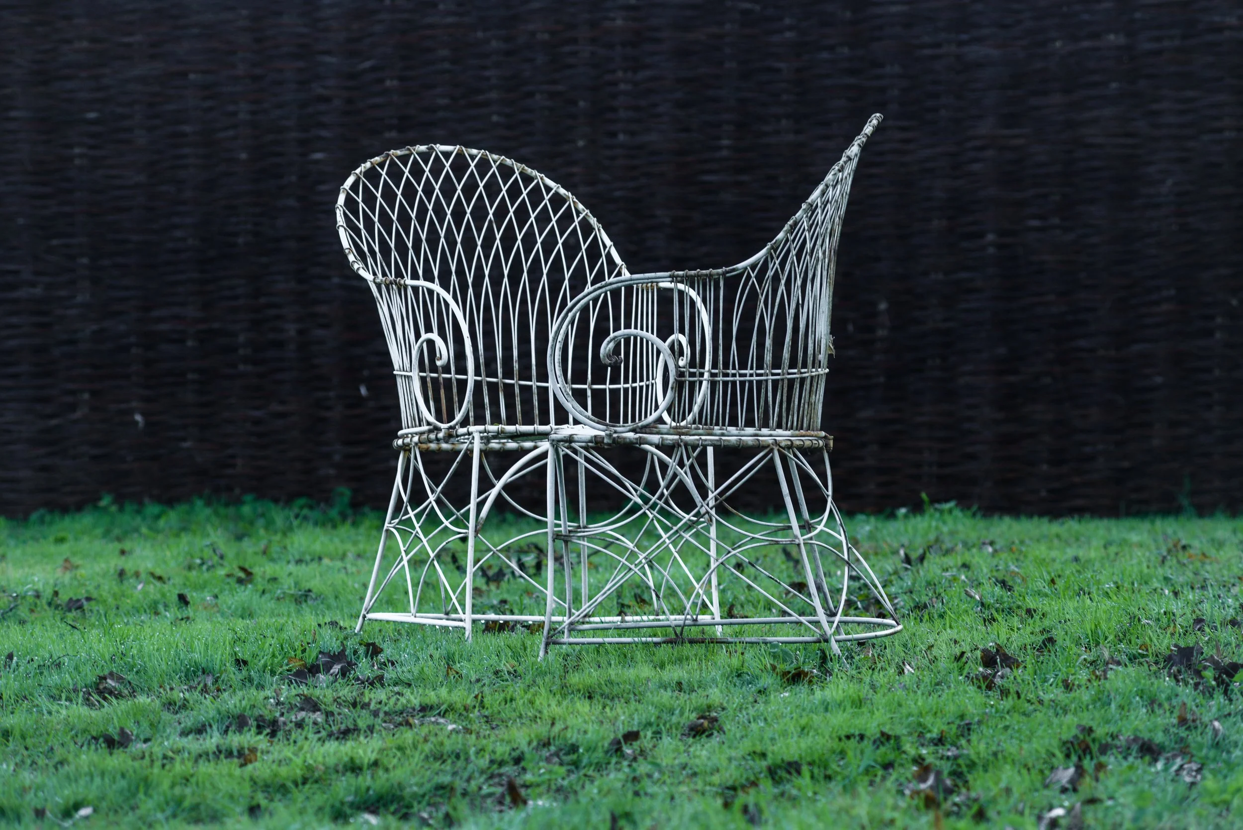 A white metal baby carriage with an ornate design sitting on green grass, with a dark fence or hedge in background.