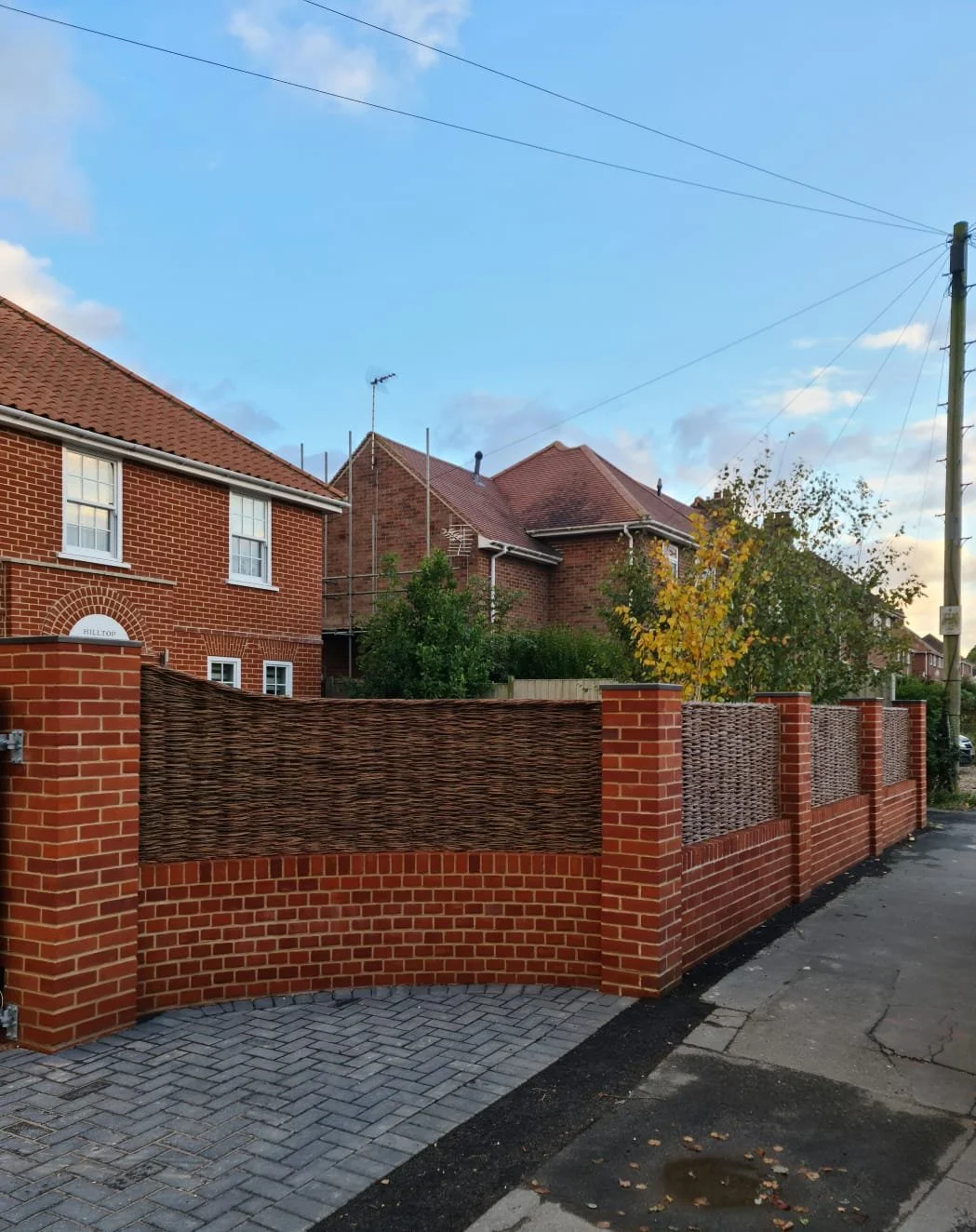 A brick residential house with a red tiled roof, surrounded by a brick and wicker-style fence, with trees and a blue sky with clouds in the background.