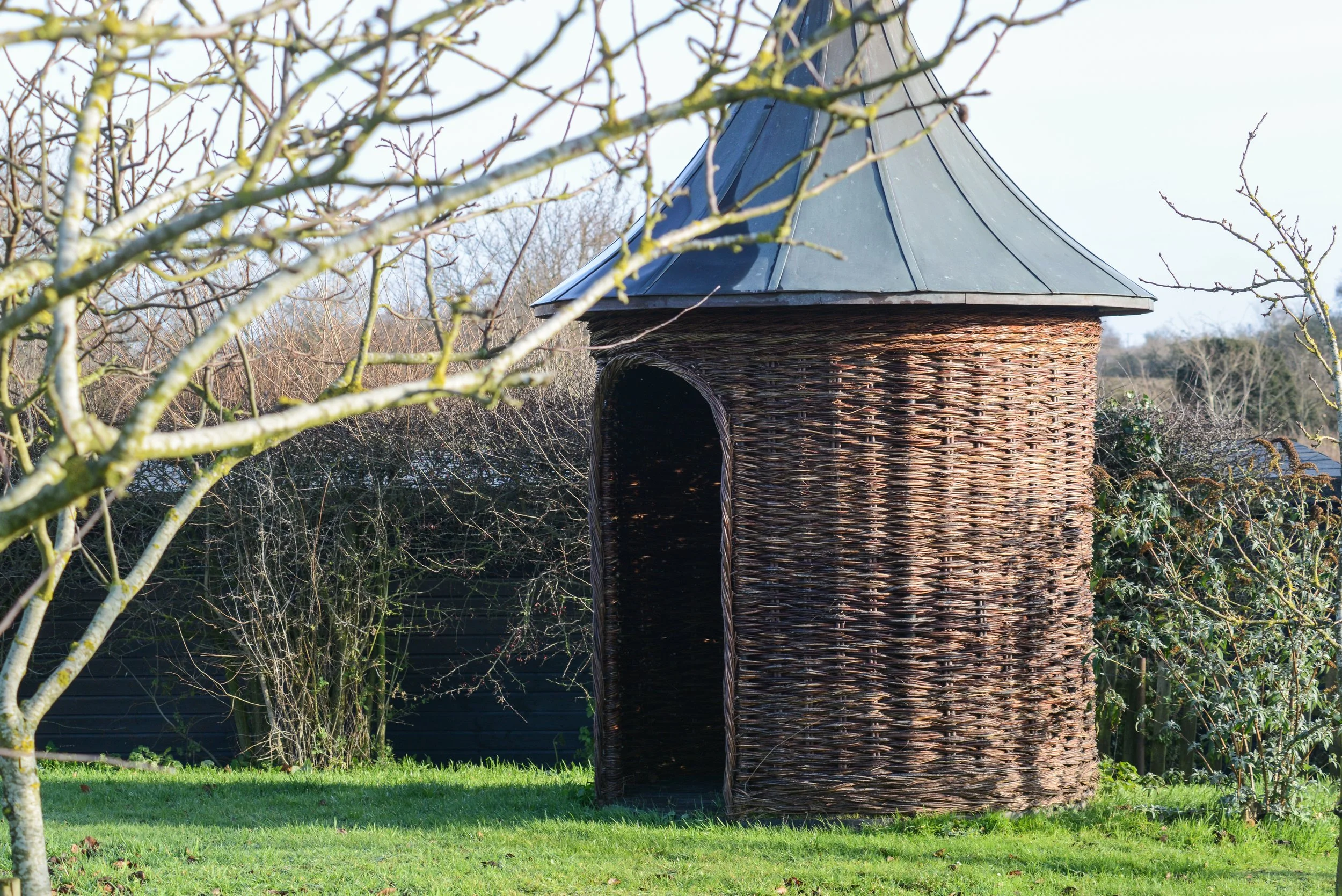 A wicker birdhouse with a metal roof set in a grassy yard, with leafless trees and bushes in the background.