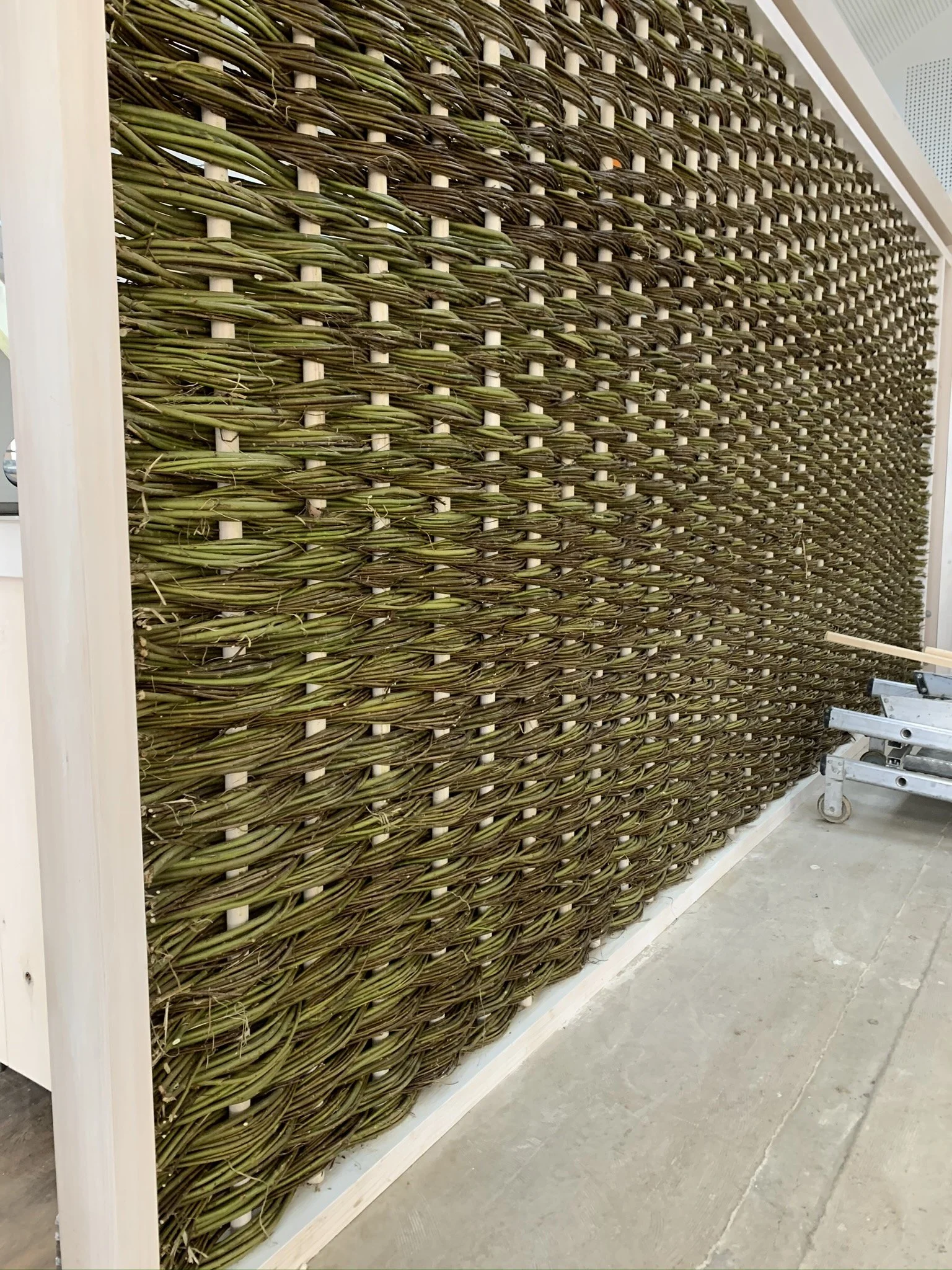 A large wall display of green and brown wicker or woven vine materials, possibly used for furniture or decoration, with part of a metal scaffold or cart nearby.