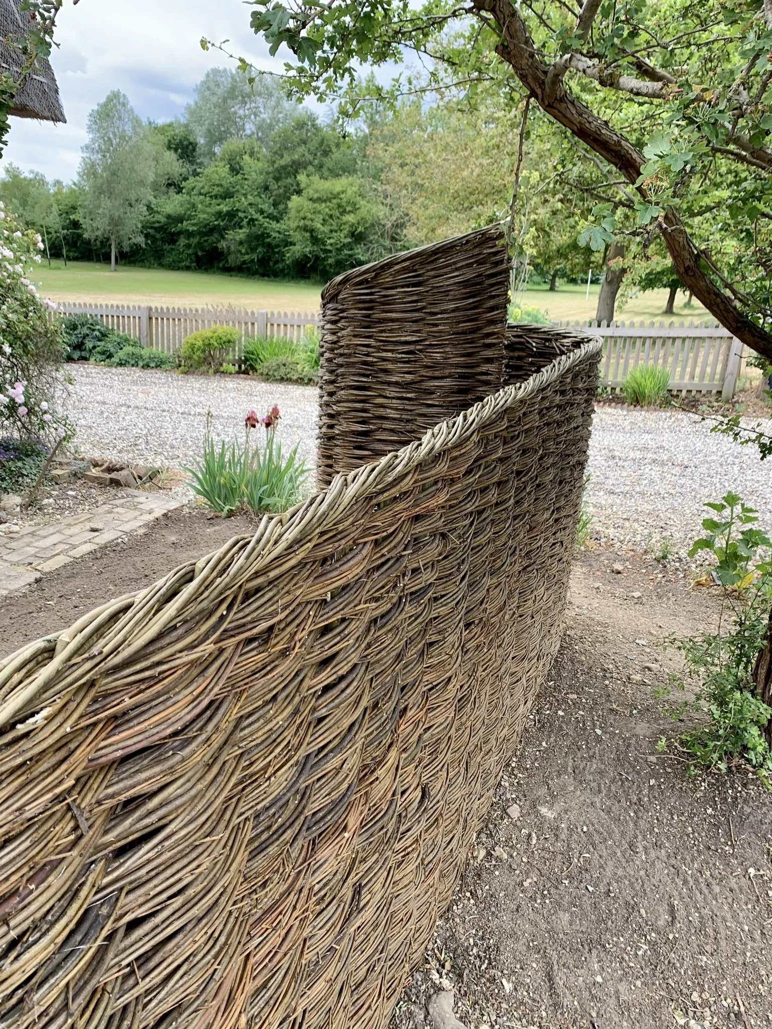 A woven wicker outdoor garden wall in a backyard garden with a gravel pathway, flowers, trees, and a wooden fence in the background.