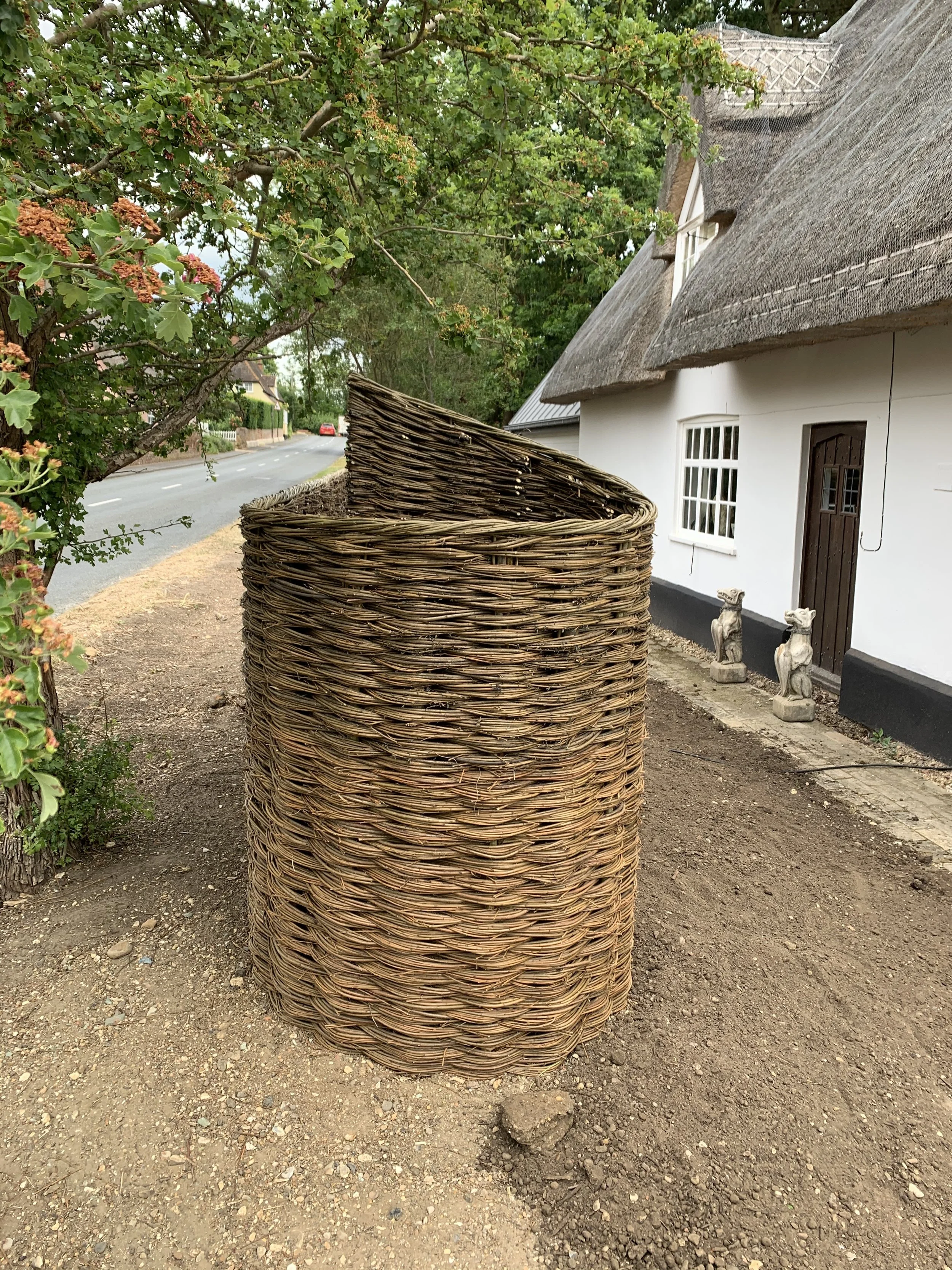 A large woven wicker basket outdoors on dirt ground next to a house with white walls and a thatched roof, with trees and a street in the background.