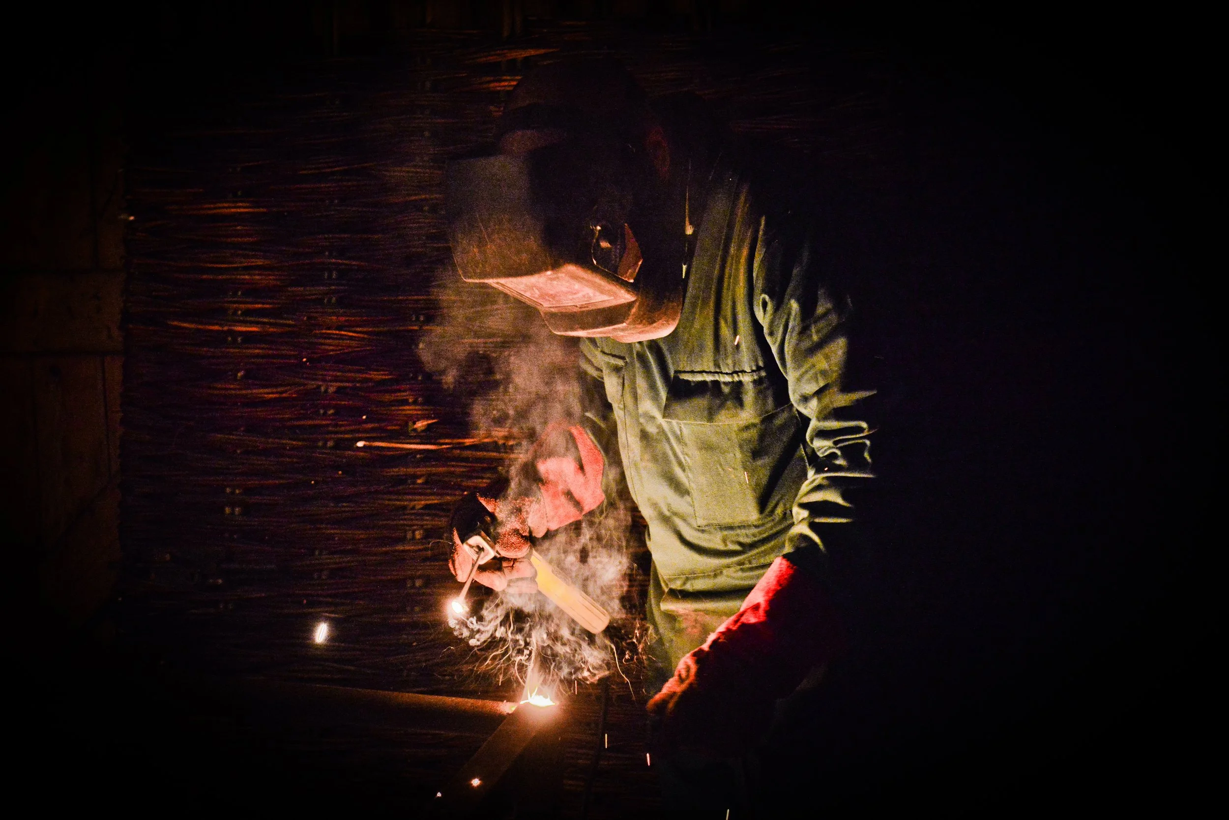 A man welding metal in a dark workshop, sparks and smoke flying as he works.