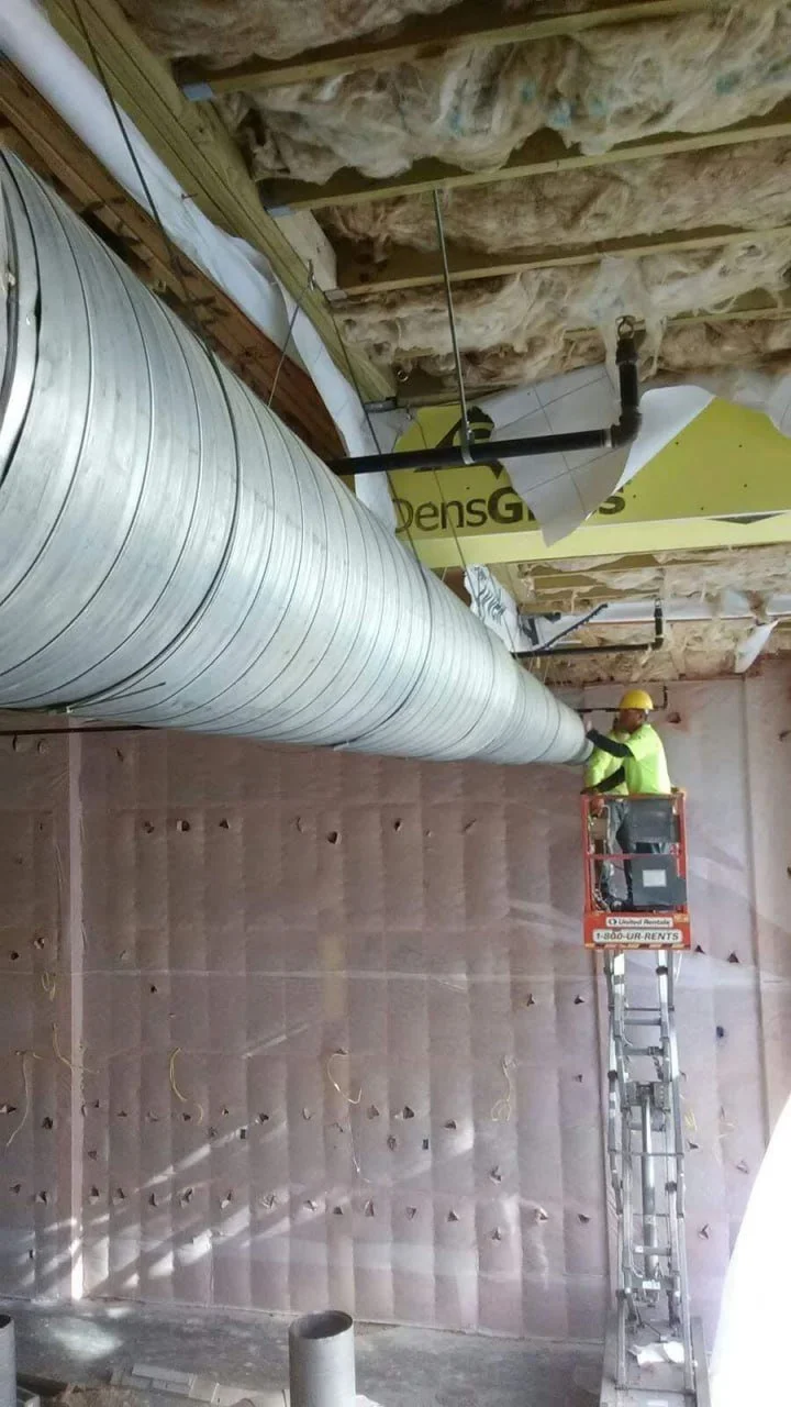 Construction worker in safety gear working on large ventilation duct inside a building under construction.