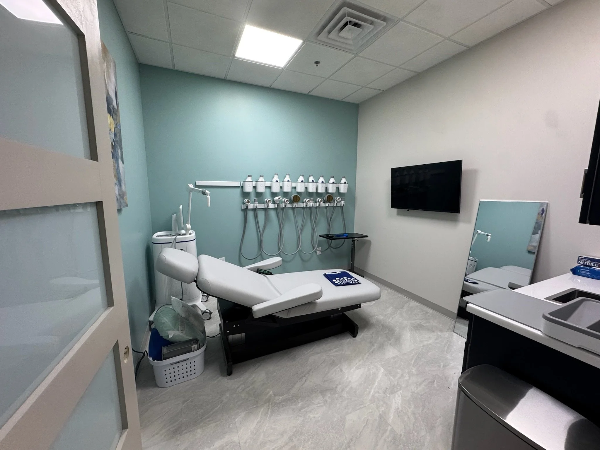 Medical examination room with a hospital bed, medical equipment, a television on the wall, and various supplies and furniture.