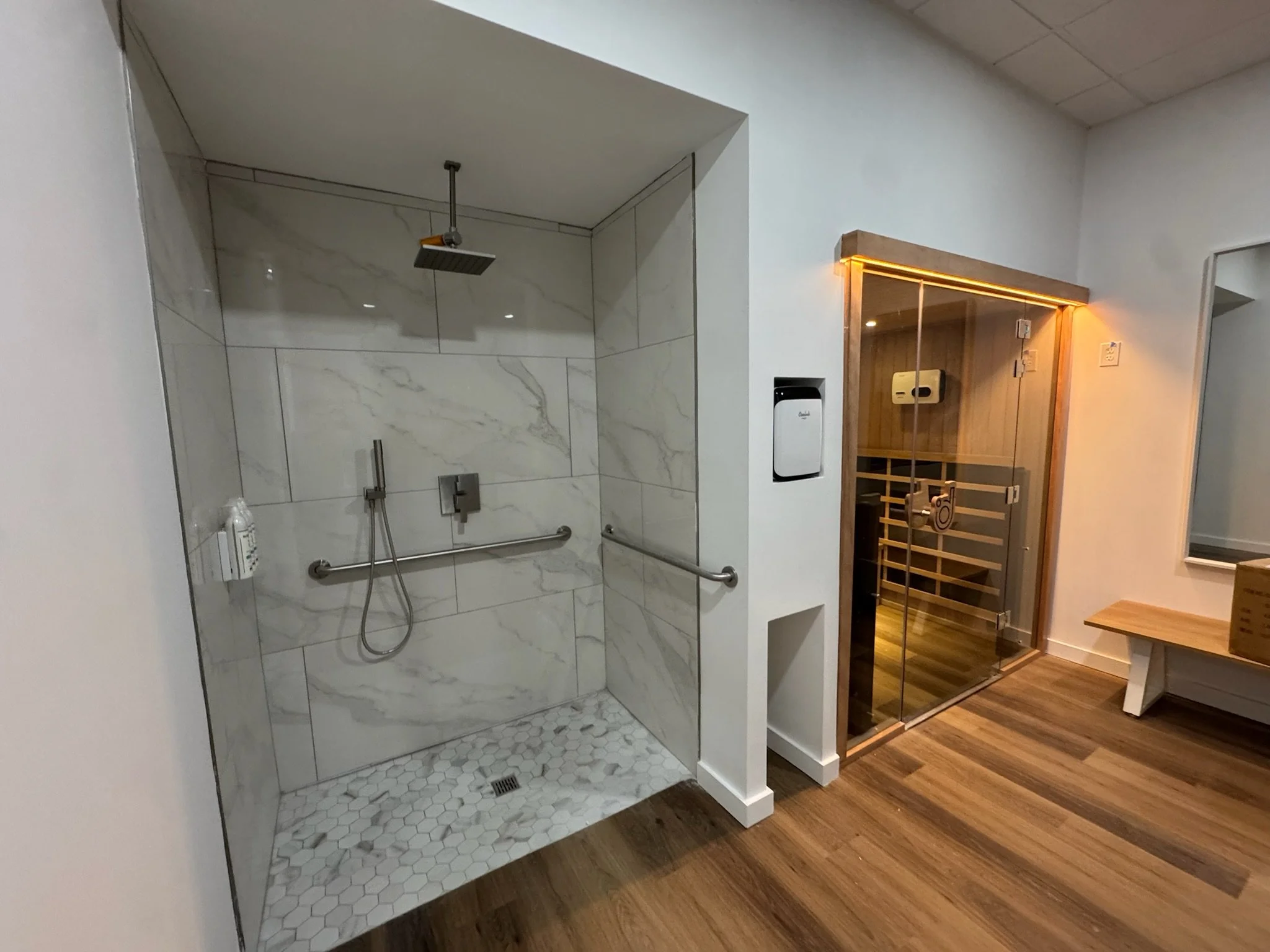 A shower area with a rainfall showerhead, grab bars, and marble-looking tiles, adjacent to a sauna with glass doors and wooden interior, located in a modern wellness or spa facility.