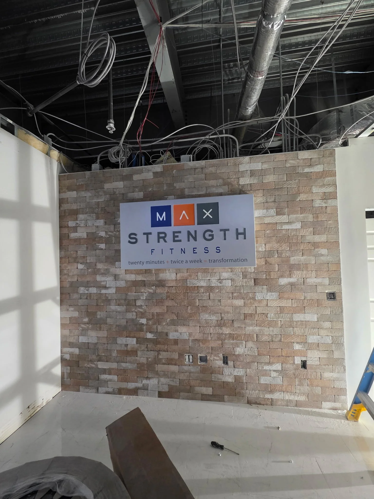 Interior of a building under construction with exposed pipes, wires, and a brick accent wall with a sign that reads 'Strength Fitness' and a slogan 'twenty minutes + twice a week = transformation'.