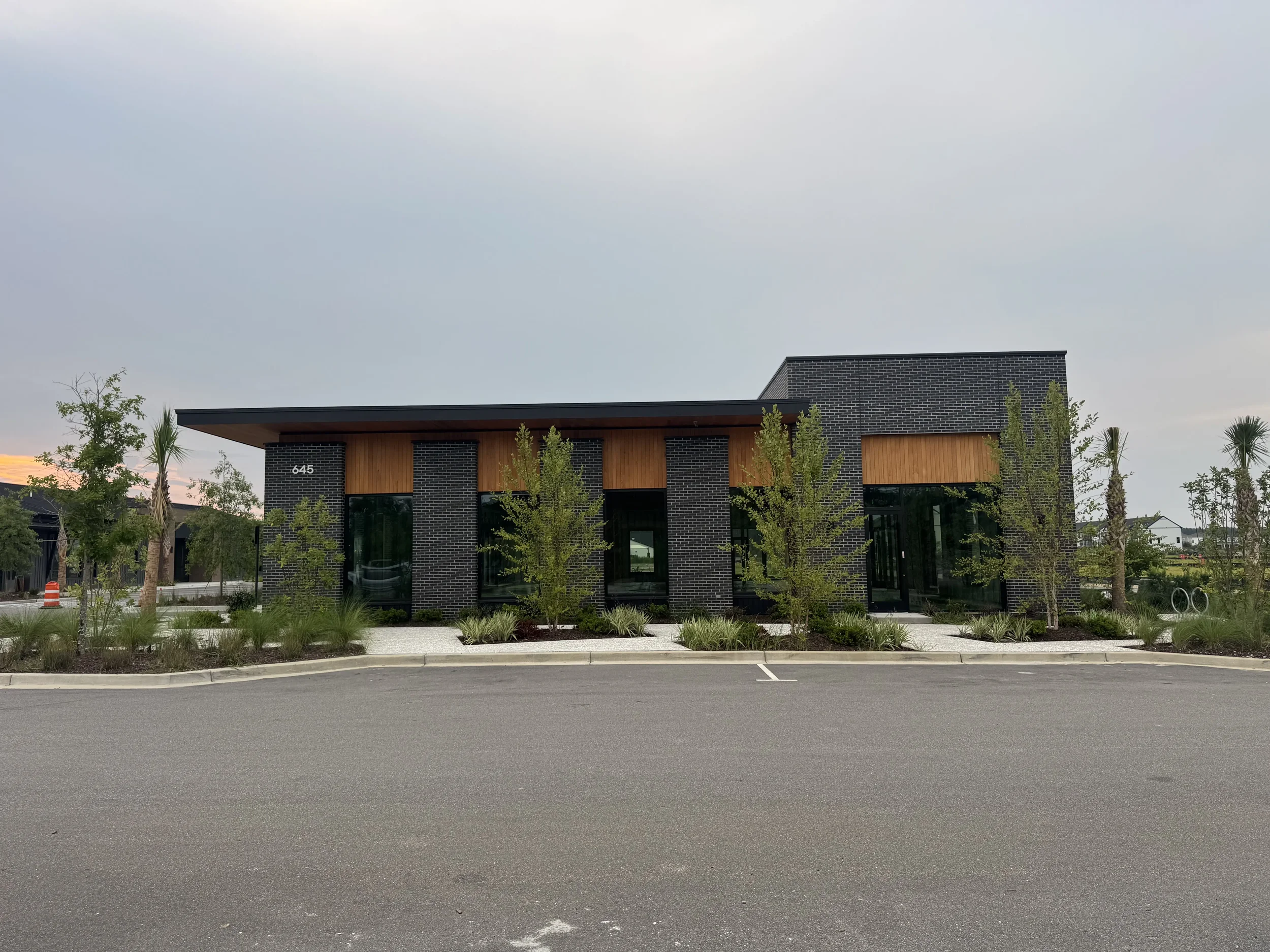 Modern building with dark brick walls, wood accents, and large windows, surrounded by young trees in a landscaped area, under a cloudy sky.