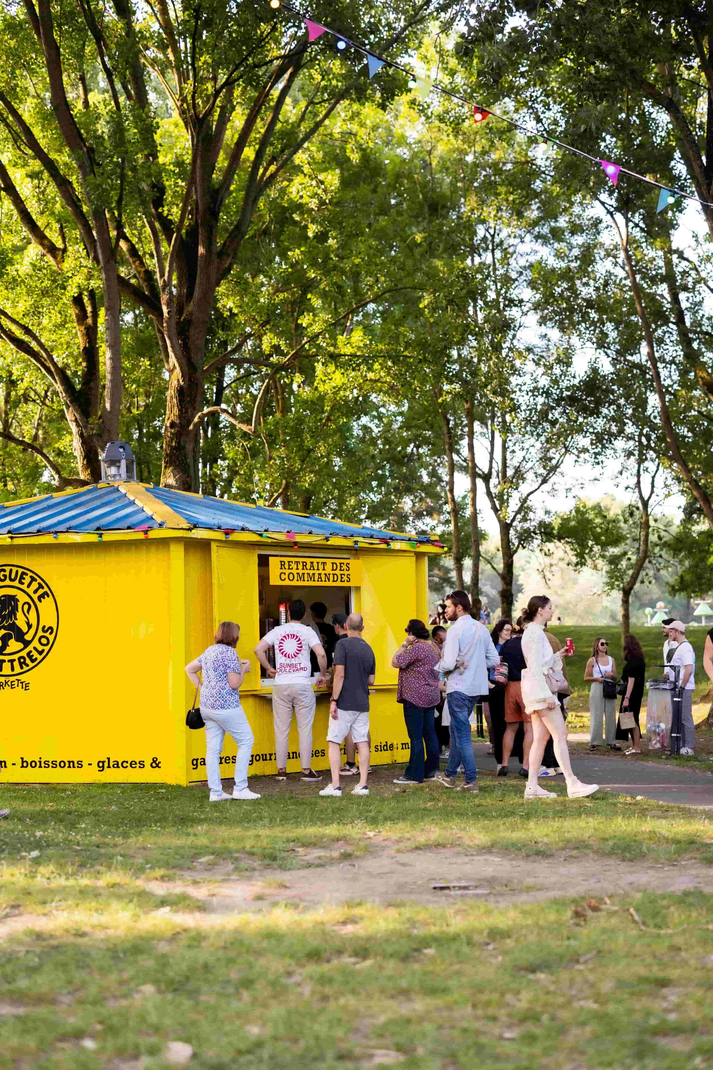 Groupe de personnes faisant la queue devant un stand jaune coloré avec l'inscription 'Prise des commandes' dans un parc, en plein air, en journée.