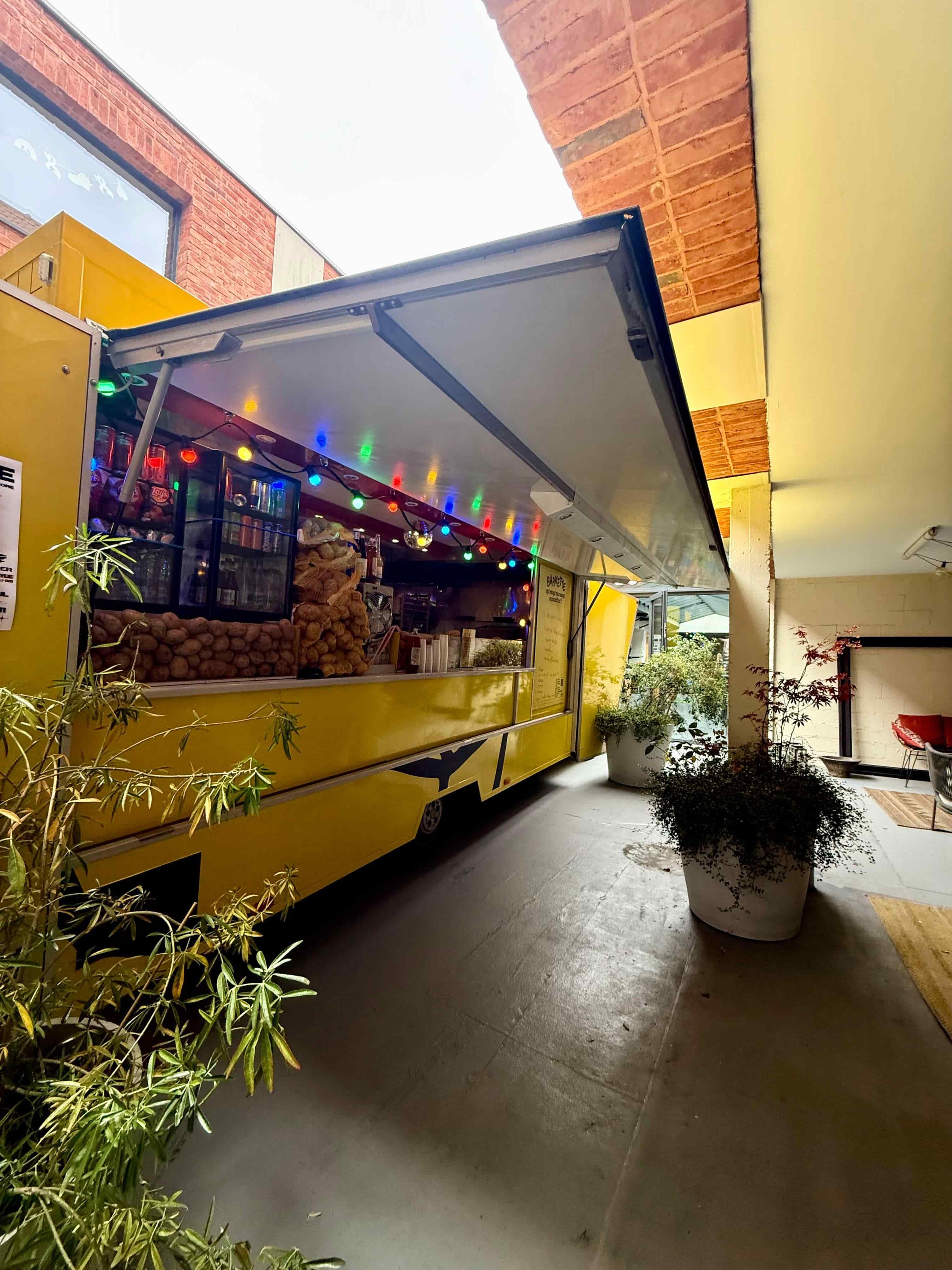 Un food truck jaune sur un plancher en intérieur, décoré avec des guirlandes de lumières colorées, entouré de plantes vertes.