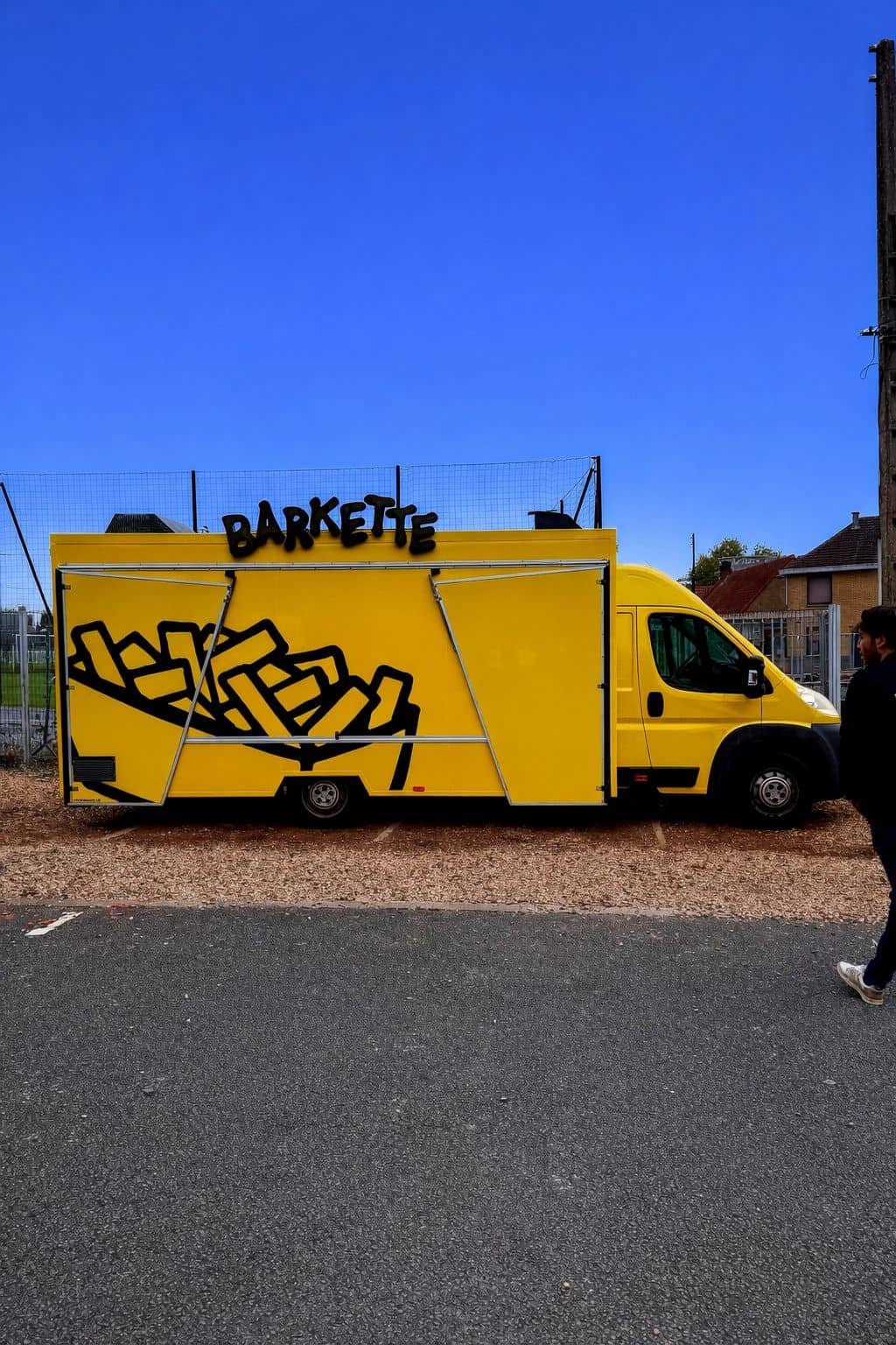 Un camion jaune avec des dessins de frites et le mot 'BARKETTE' en haut, stationné à côté d'un terrain de sport et d'un bâtiment résidentiel, sous un ciel bleu clair.
