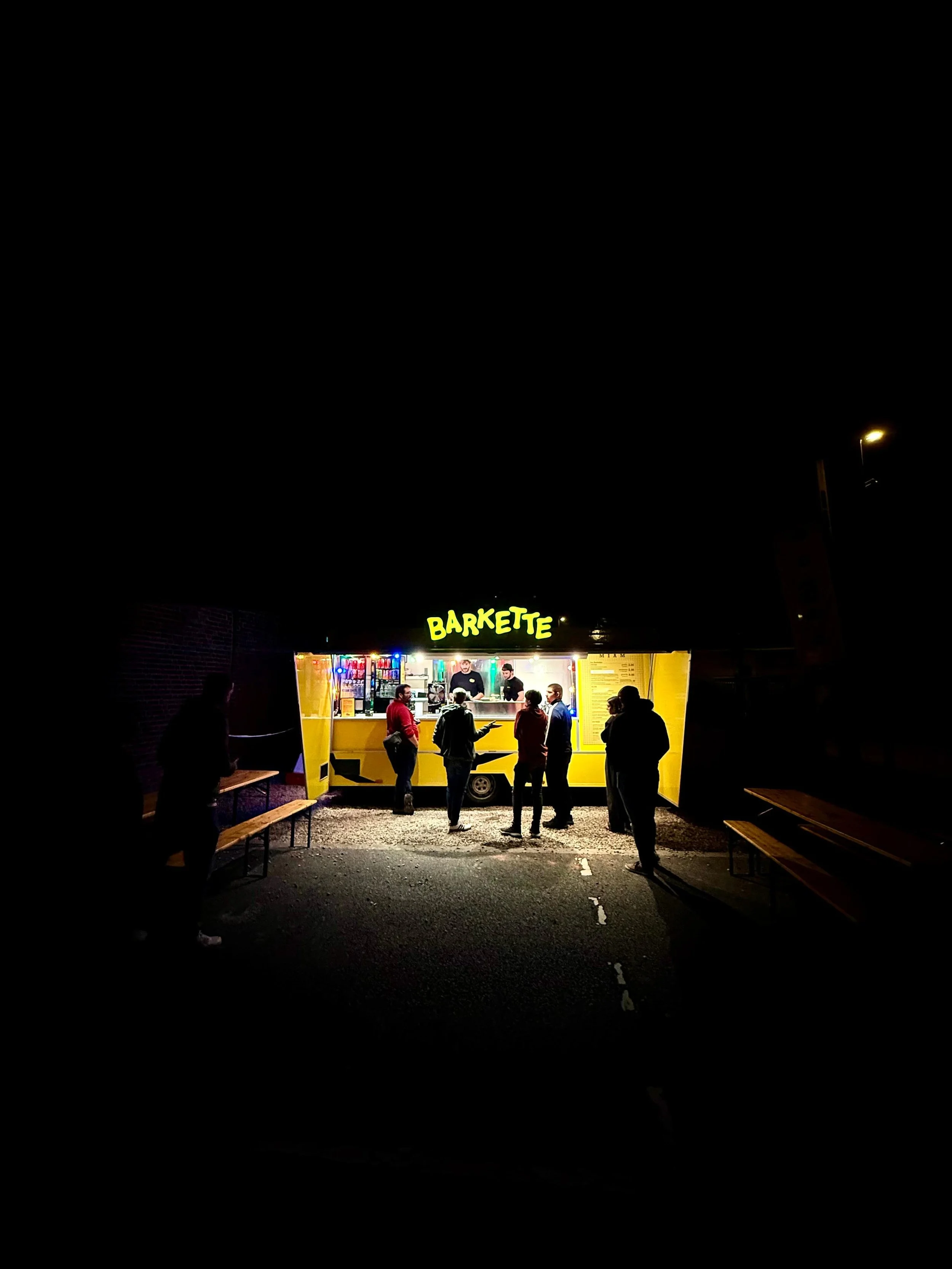 Un stand de nourriture lumineux jaune, nommé 'BARKETTE', avec des gens qui achètent et attendent leur commande, la nuit.