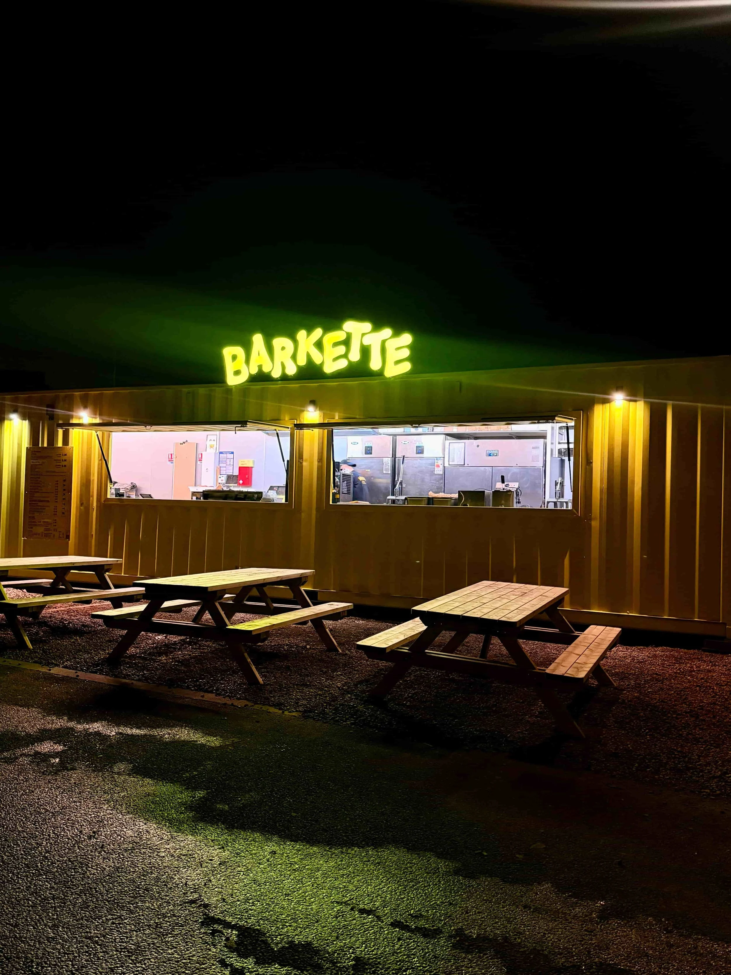 Restaurant appelé 'Barkette' avec un comptoir visible à l'intérieur, éclairé la nuit, avec plusieurs tables en bois devant.
