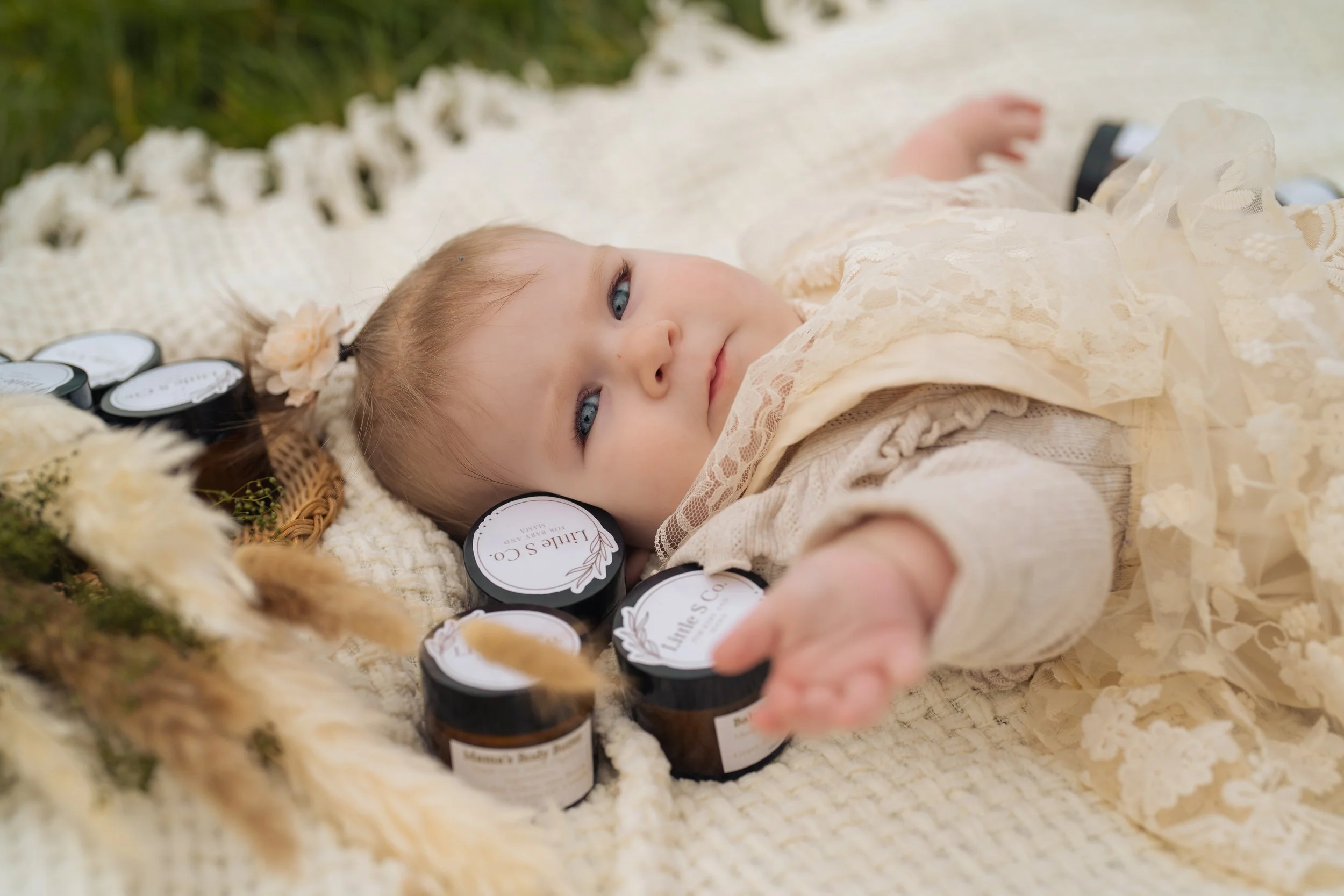 A young girl lying on a blanket outdoors, surrounded by jars labeled "Mama's Body Butter" by Little S Co. and other natural skincare products, with a floral headband, dressed in a lace dress, reaching out with her hand.