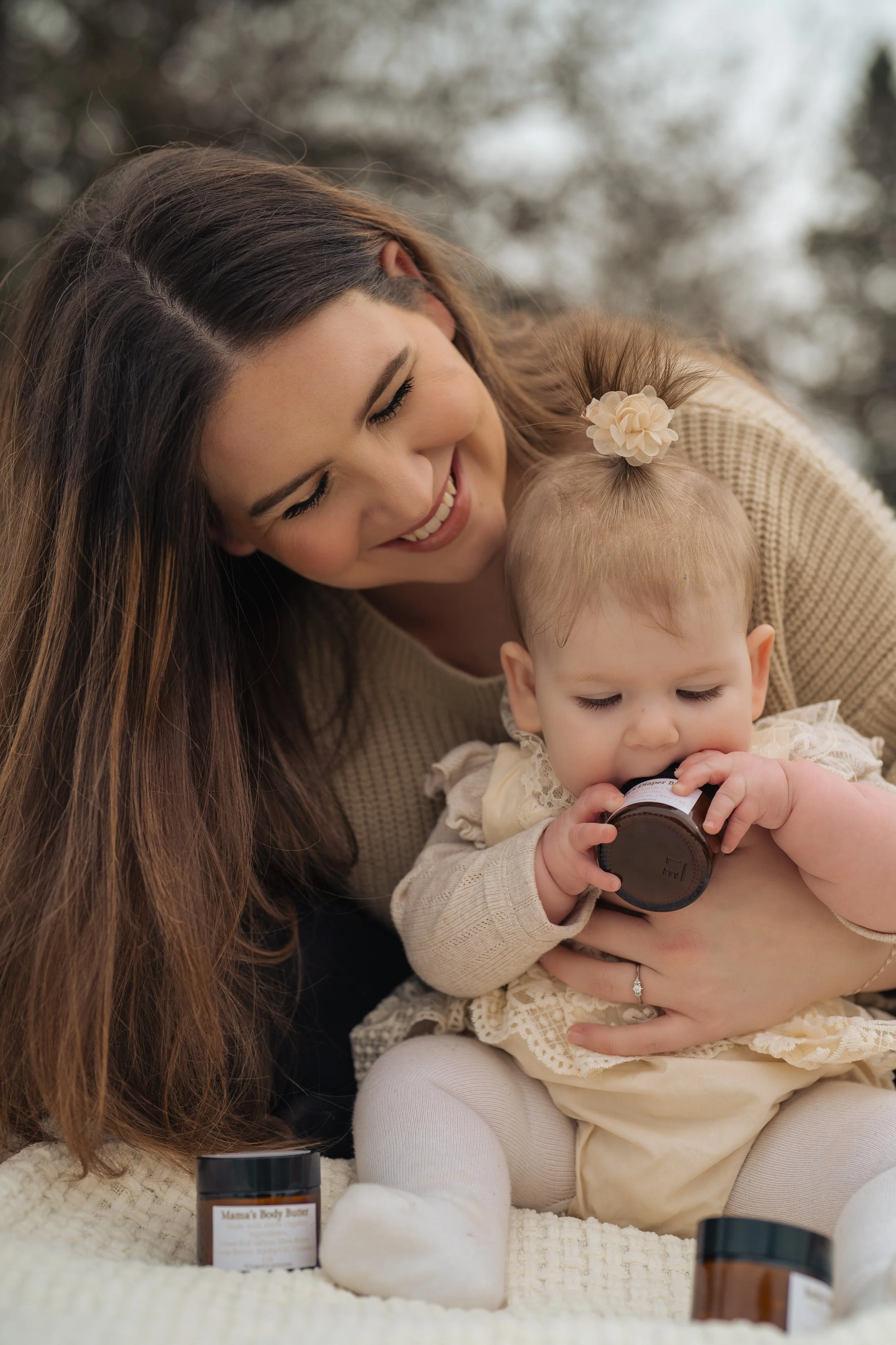 A woman with long brown hair smiling and cuddling a baby girl who is holding tallow-based skincare product by Little S Co. The baby has a flower hair clip and is dressed in beige. There are more tallow-based skincare products in front of them.